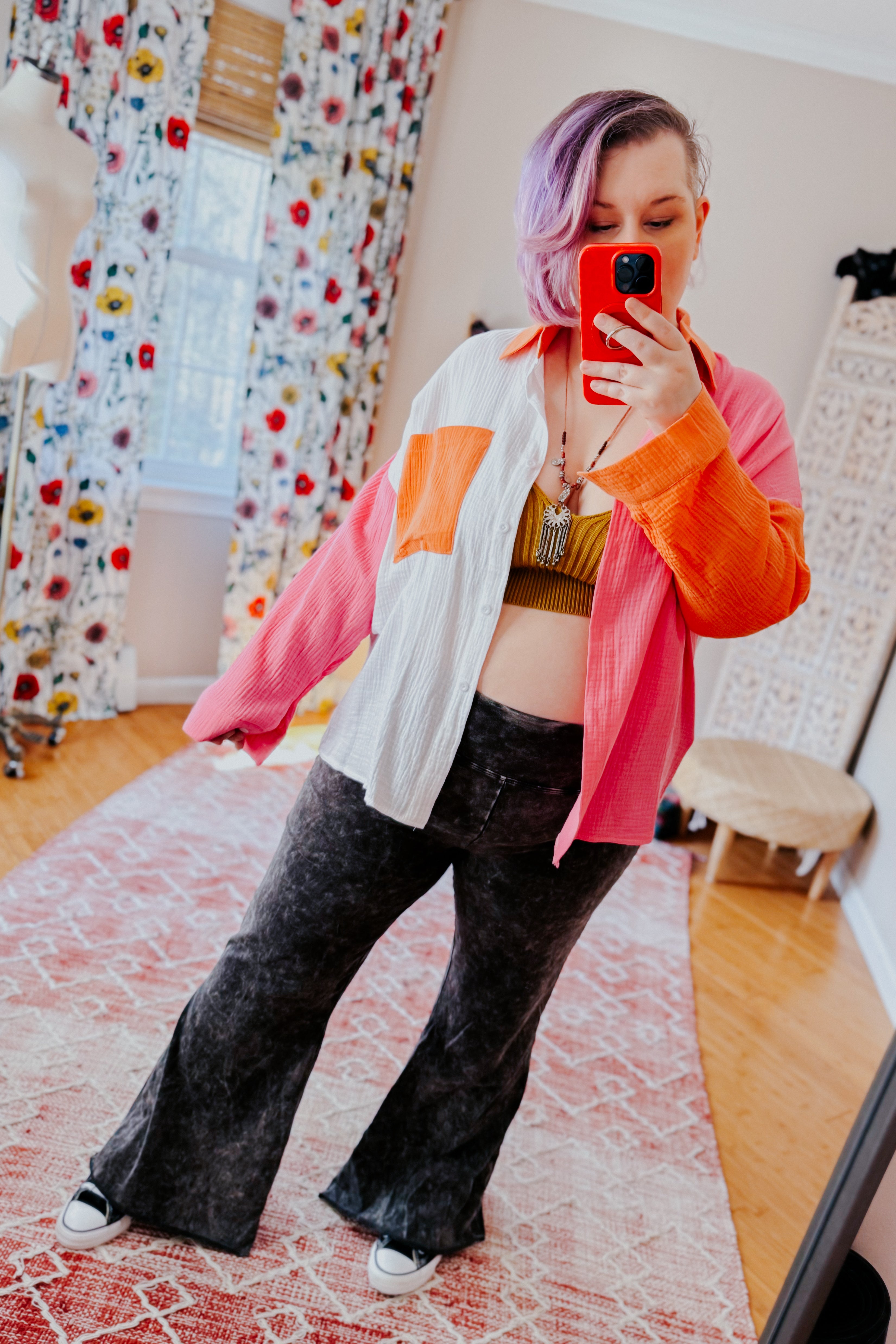 Woman with purple hair wearing pink, orange, and ivory colorblock shirt over green crop top and black flared pants taking mirror selfie in room