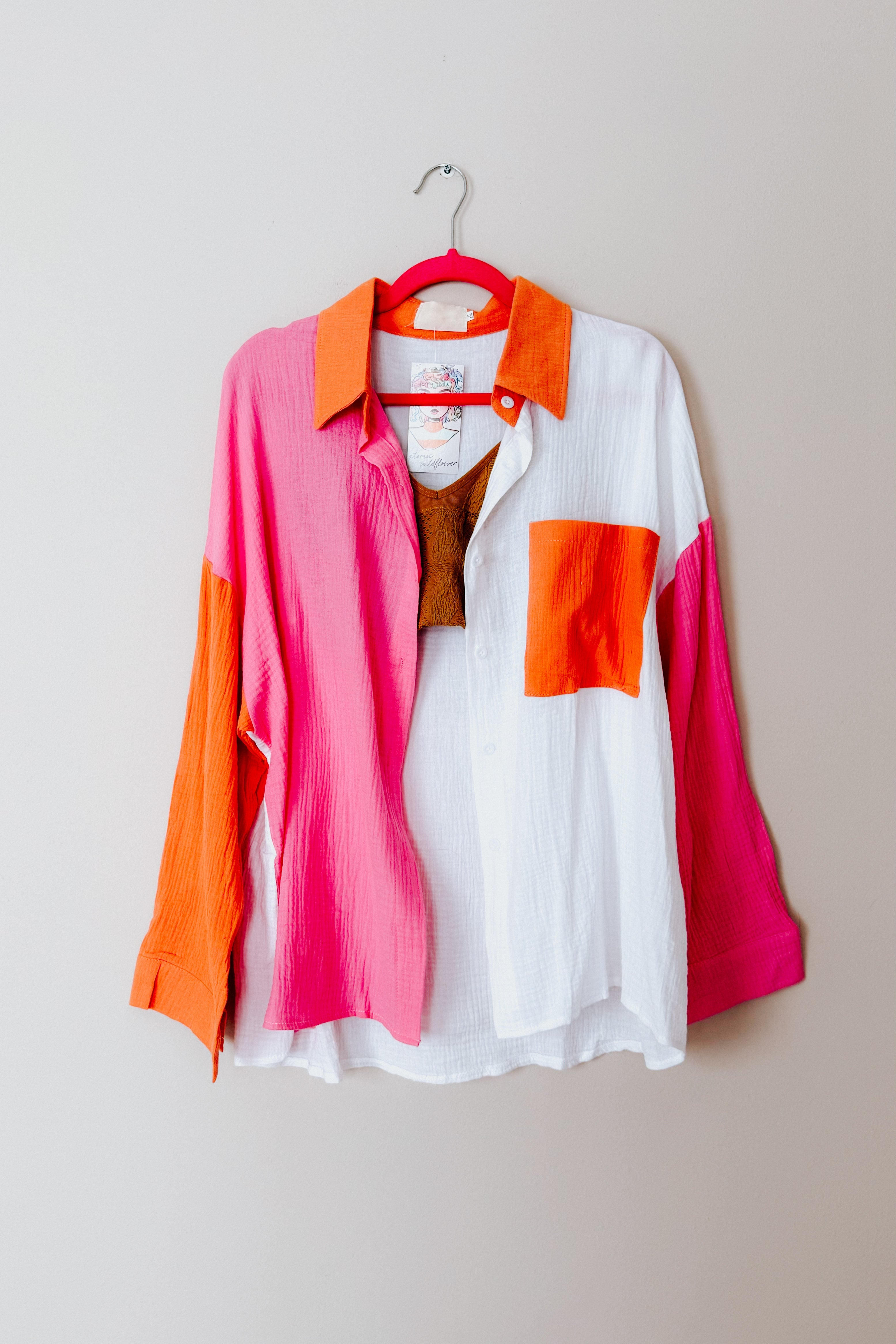Lightweight button-up shirt with pink, orange, and ivory color blocks hanging on a bright pink hanger against a plain wall