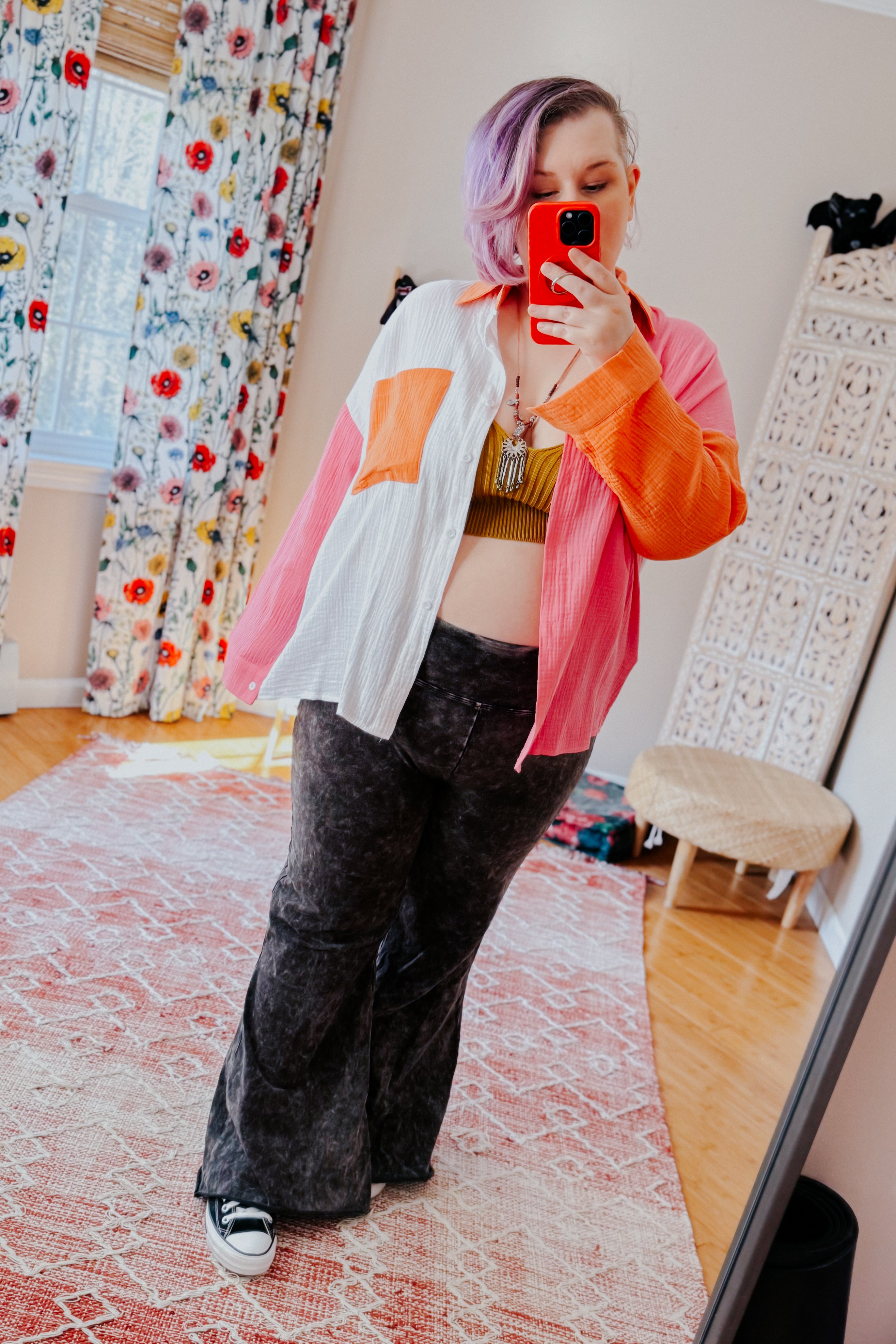 Woman with lavender hair wearing a pink, orange, and ivory colorblock shirt over olive crop top and black pants taking selfie indoors