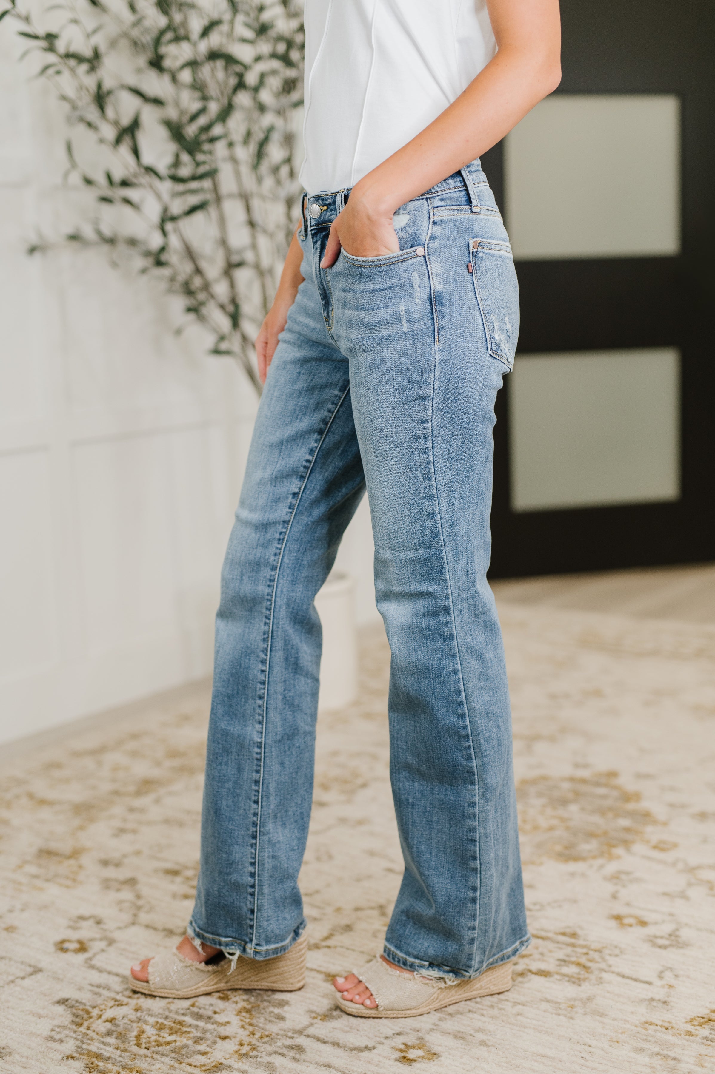 Side view of mid-rise blue denim jeans with slight fading and raw hem paired with beige wedge sandals indoors