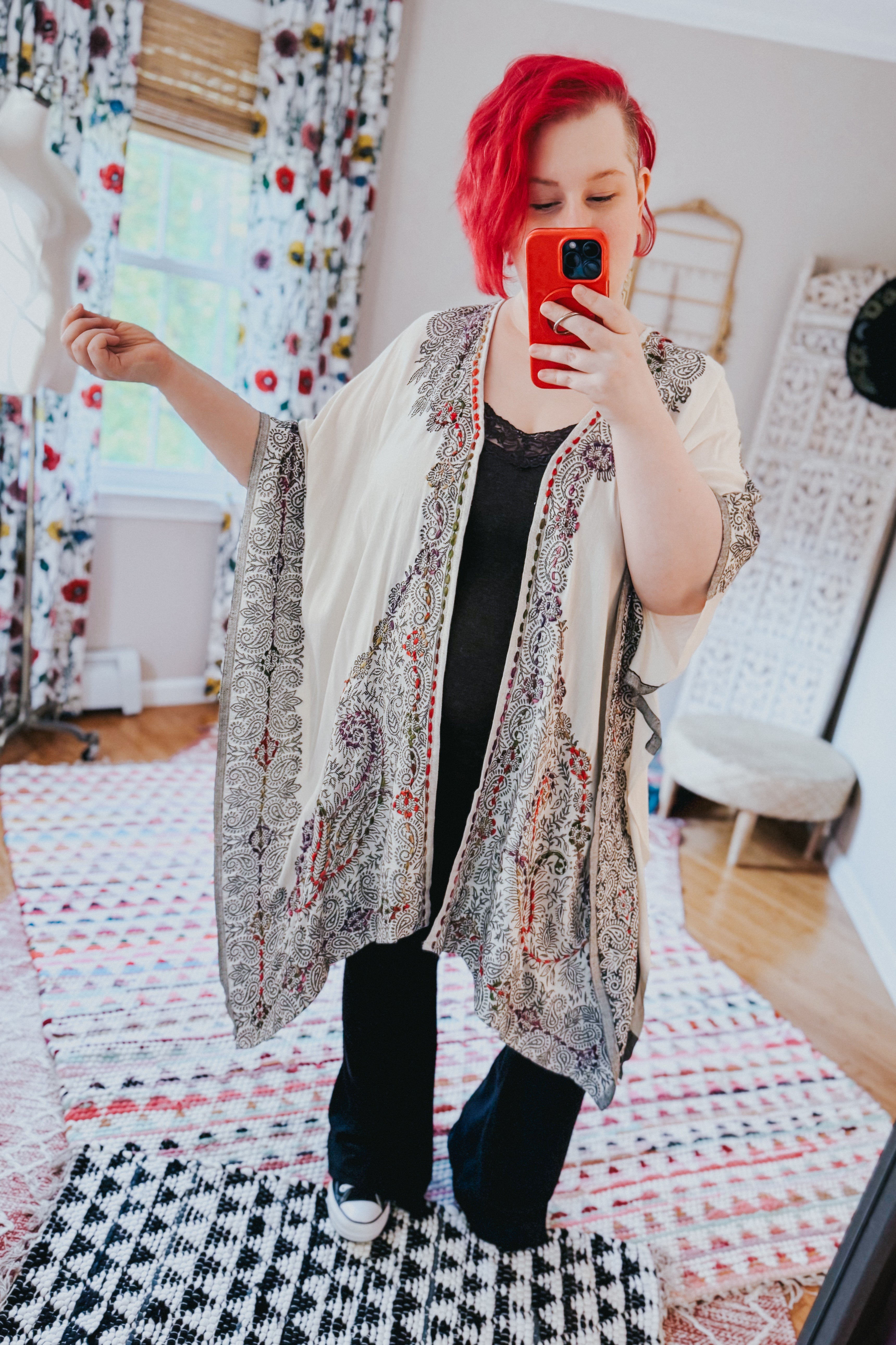 Woman wearing ivory and black patterned duster over black top and pants taking indoor mirror selfie in bright room