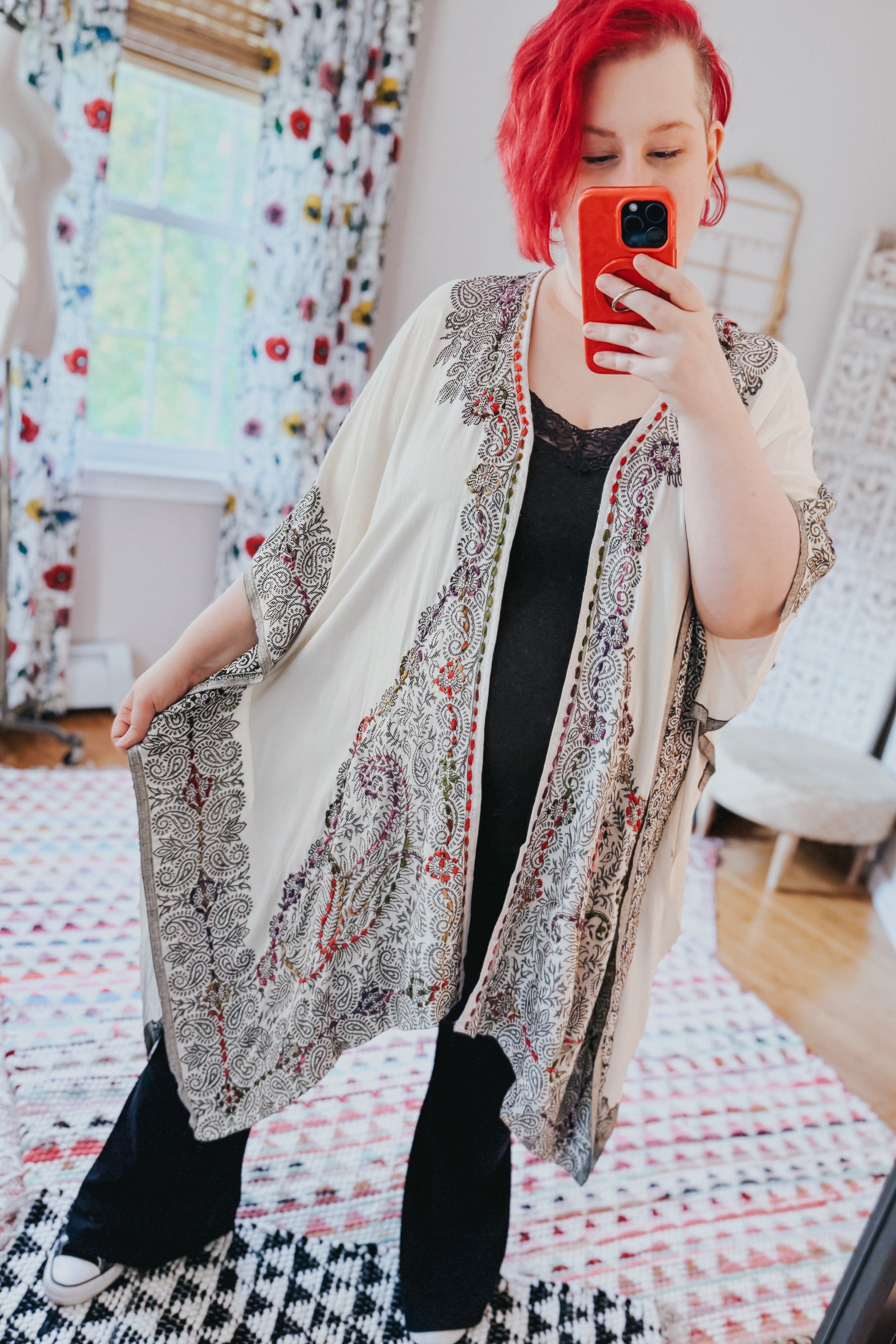 Woman with red hair taking selfie wearing ivory and black patterned duster over black lace top and pants indoors