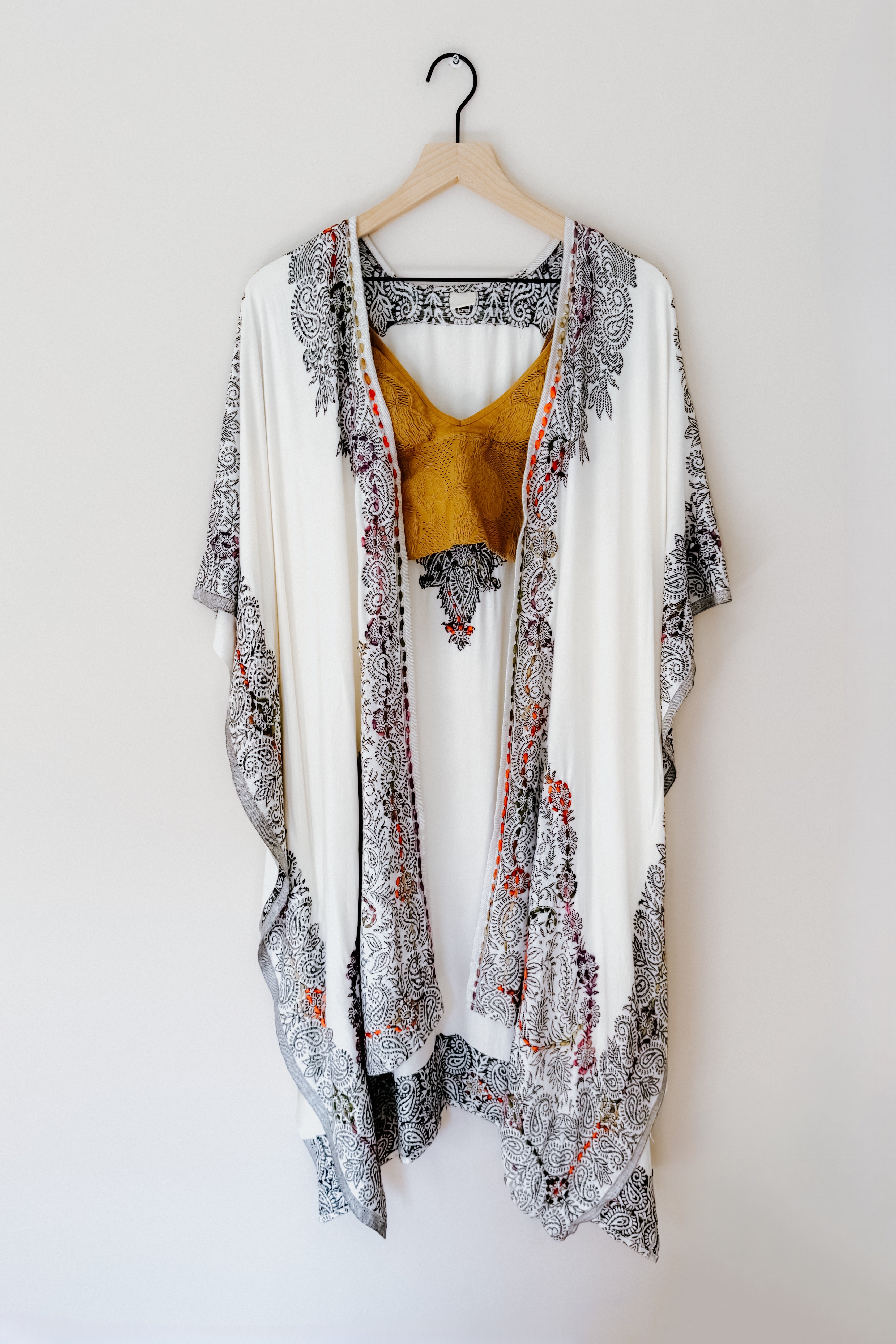 Ivory and black patterned duster layered over a mustard textured sleeveless top hanging on wooden hanger against white wall