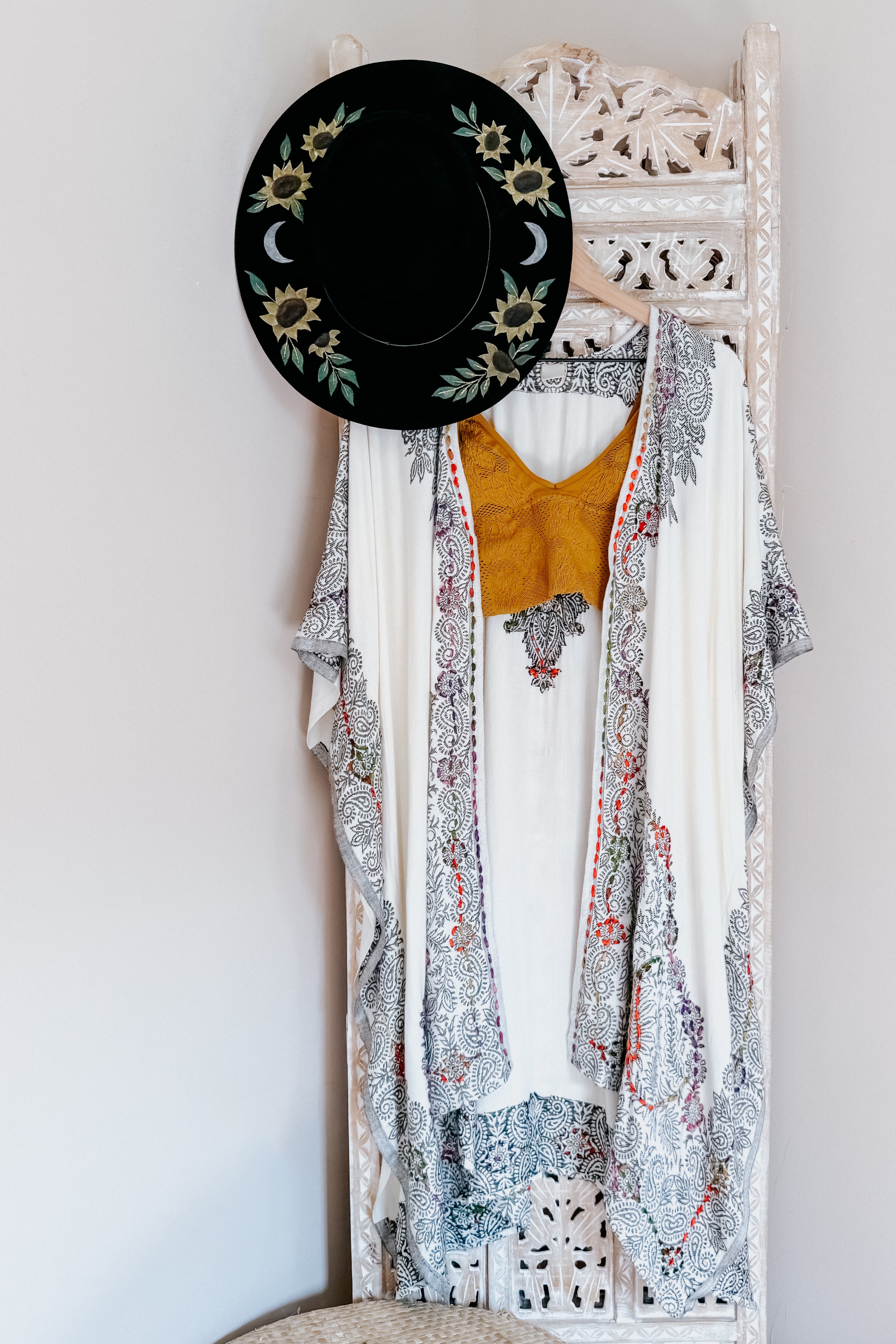 Ivory and black patterned duster layered over mustard textured top hanging on carved wooden screen with black hat adorned with sunflowers and moons
