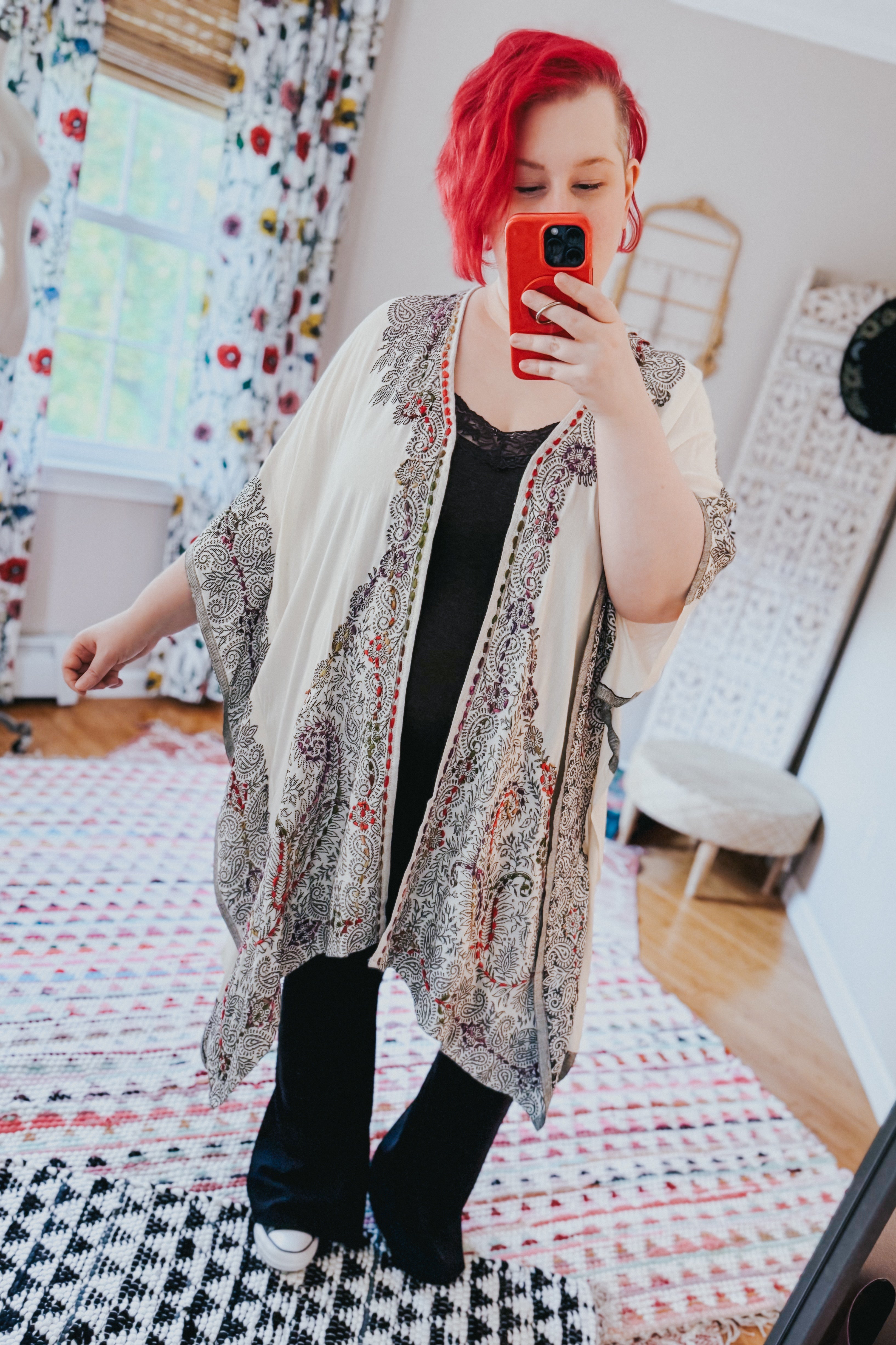 Woman with red hair wearing ivory and black patterned duster over black lace top and pants taking selfie indoors
