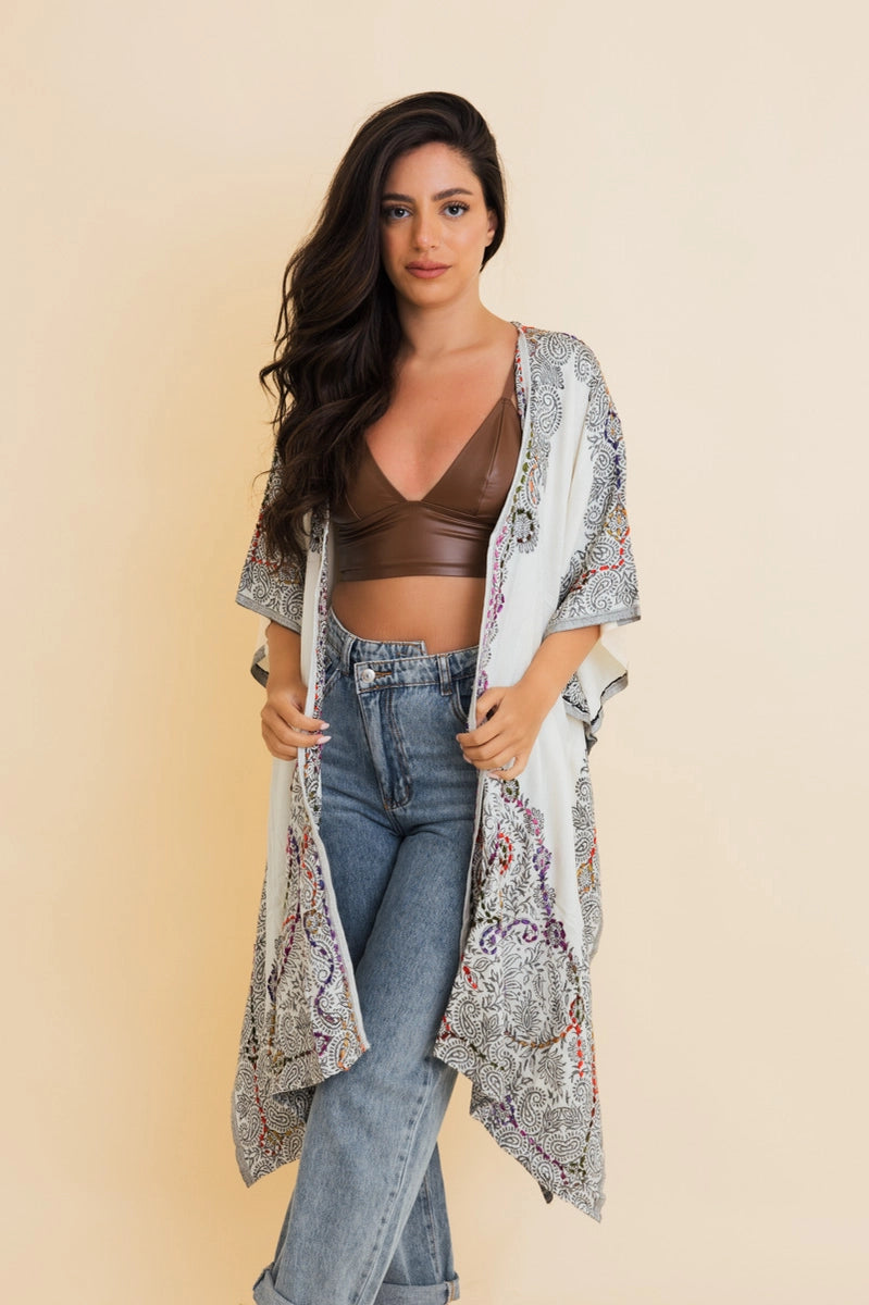 Woman wearing ivory and black patterned duster over brown bralette and high-waisted light blue jeans standing against beige background
