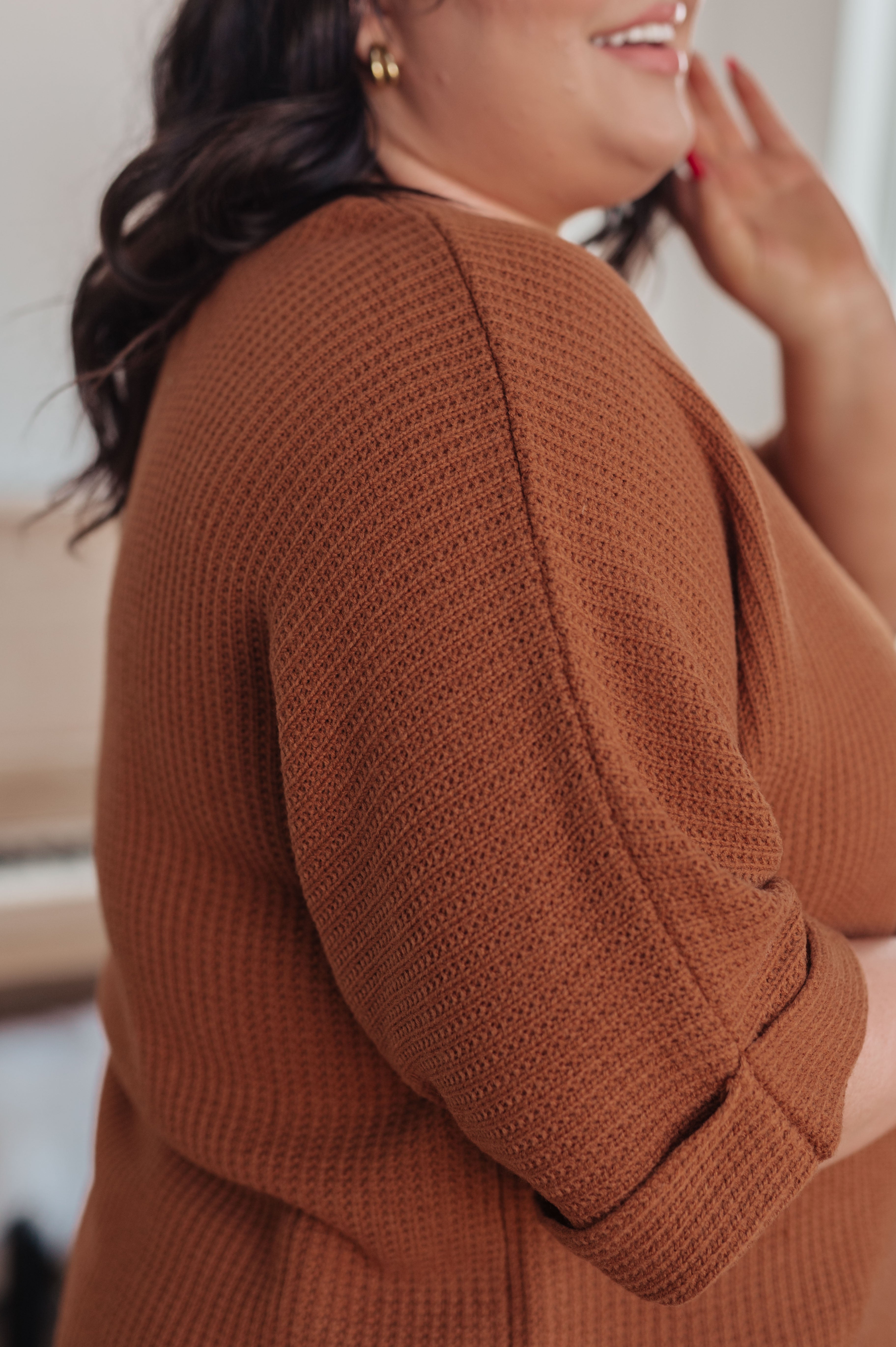 Curvy woman wearing a textured rust-colored knit top with rolled sleeves smiling indoors with hand near face