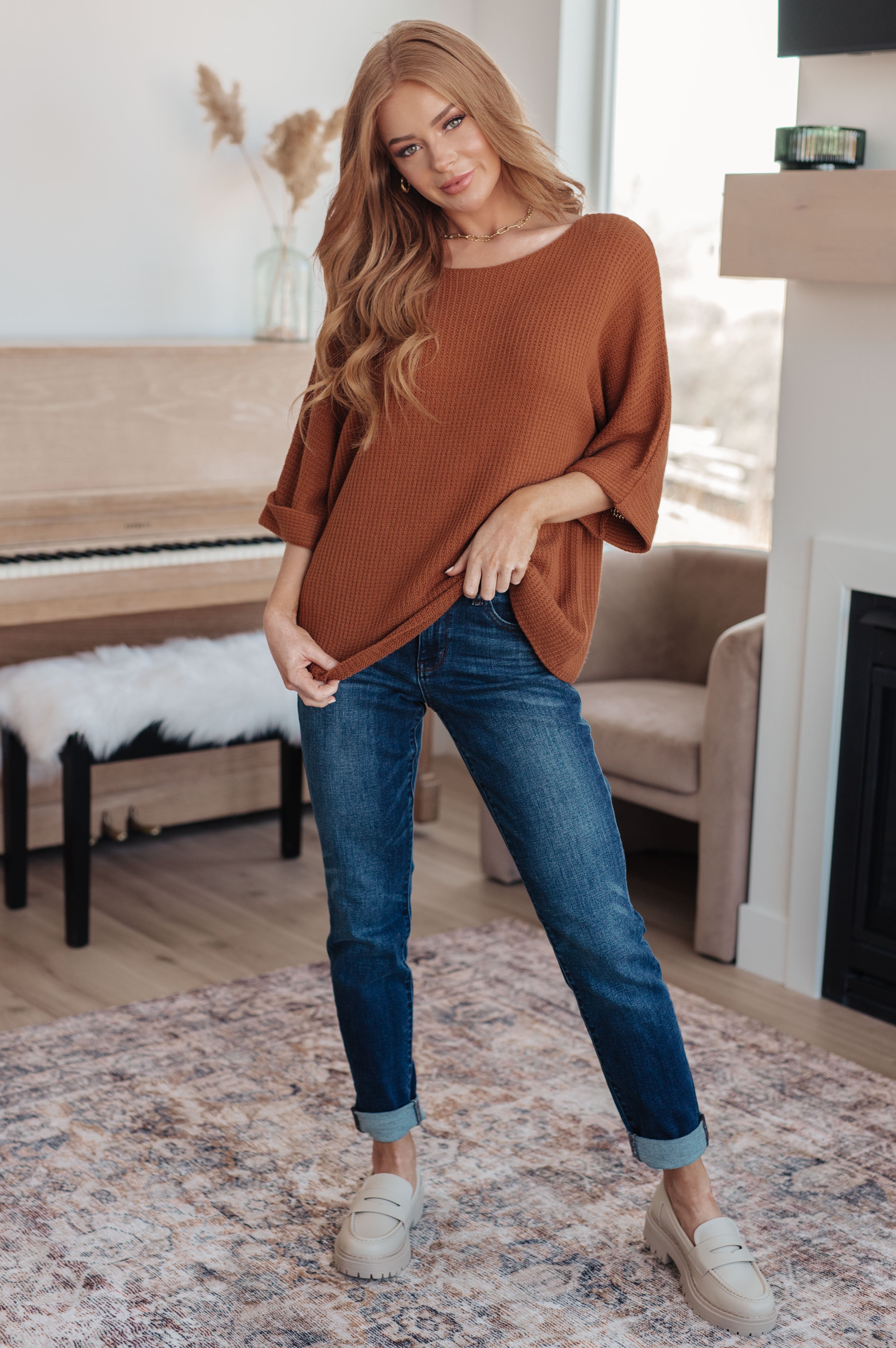 Young woman indoors wearing relaxed rust-colored knit top with rolled sleeves and cuffed dark blue jeans standing by piano