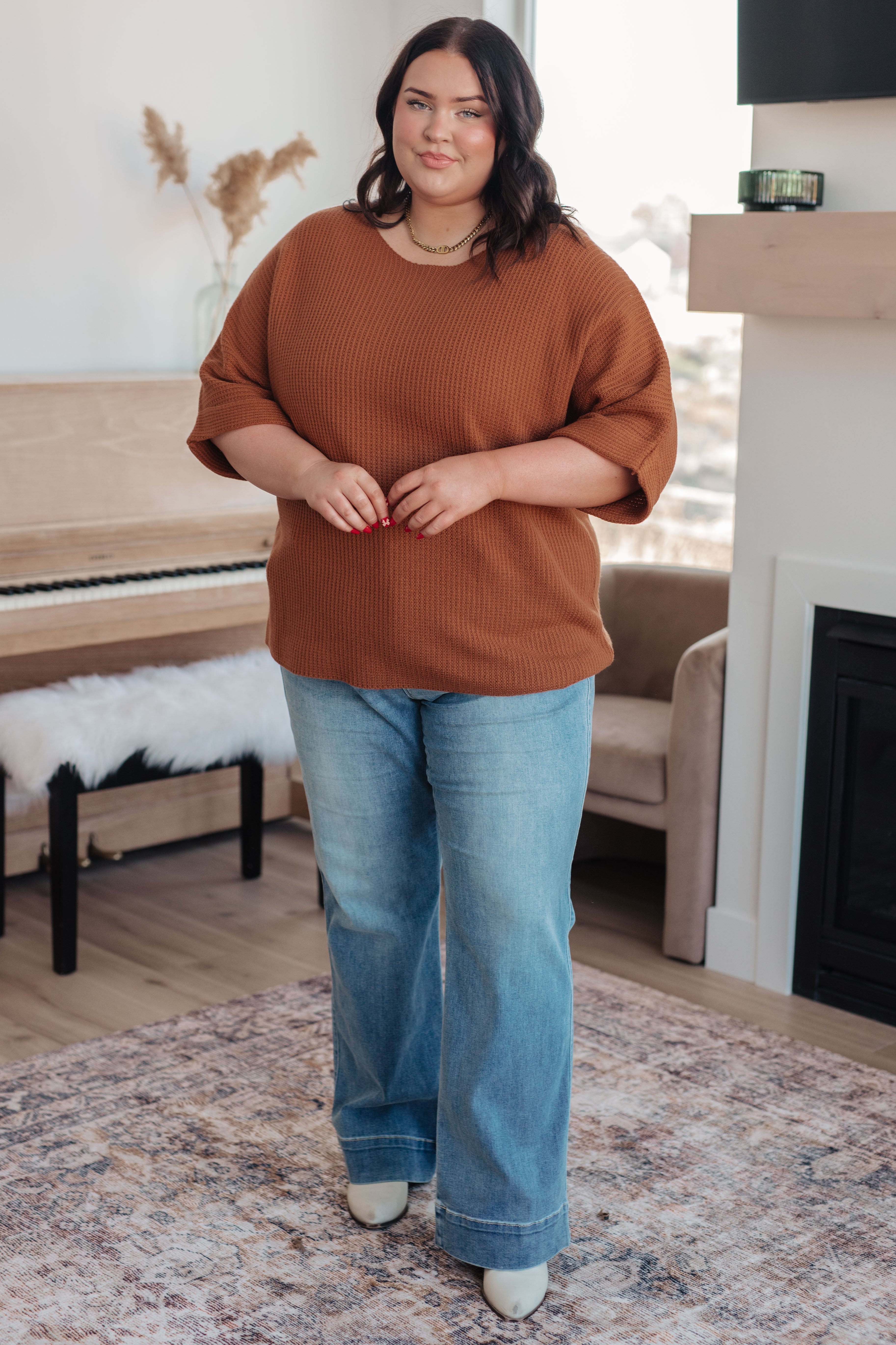 Curvy woman wearing rust-colored ribbed knit top paired with wide-leg light blue jeans standing indoors near piano
