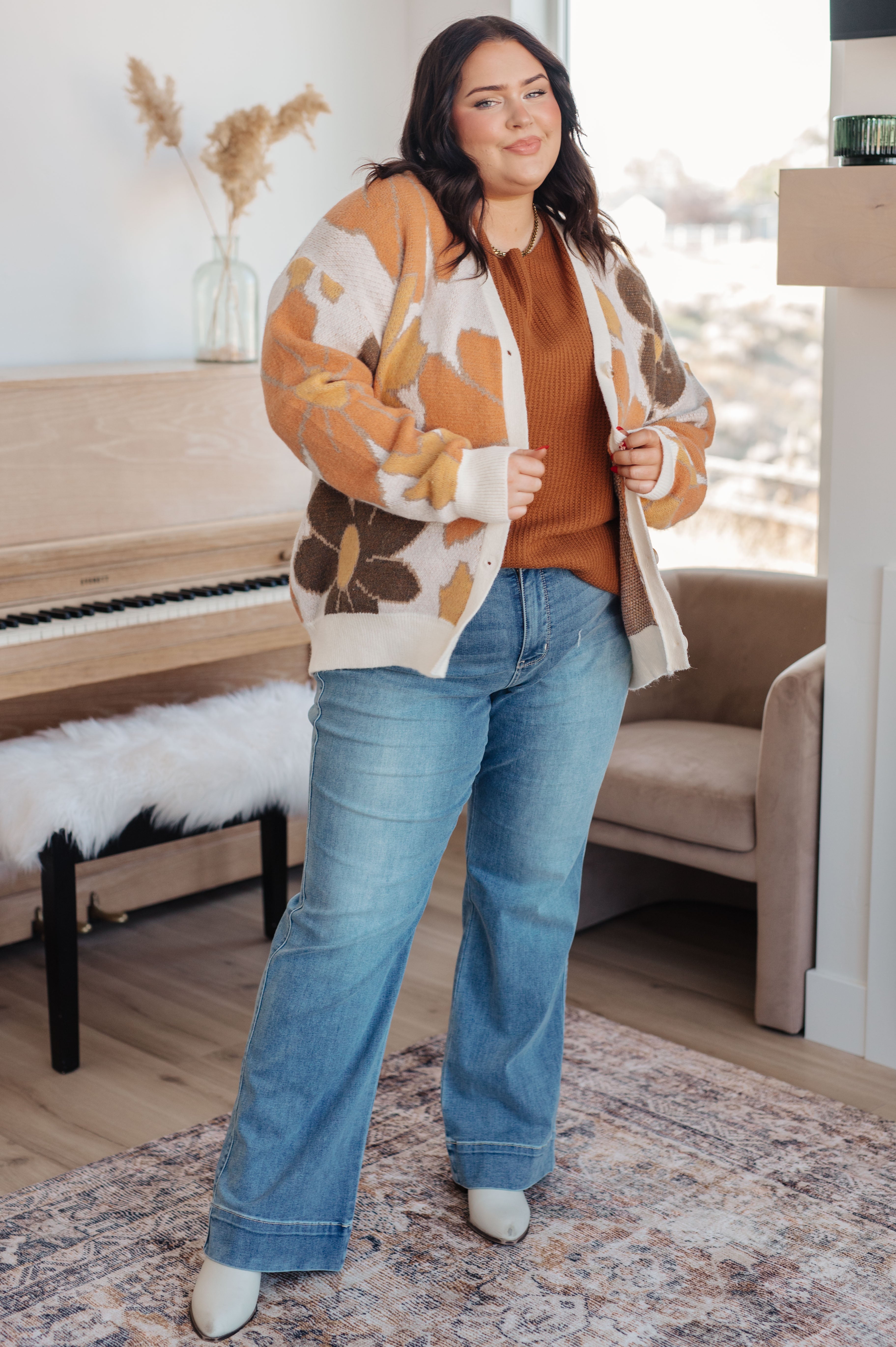 Curvy woman wearing rust-colored sweater paired with floral patterned cardigan and light blue wide-leg jeans indoors by piano