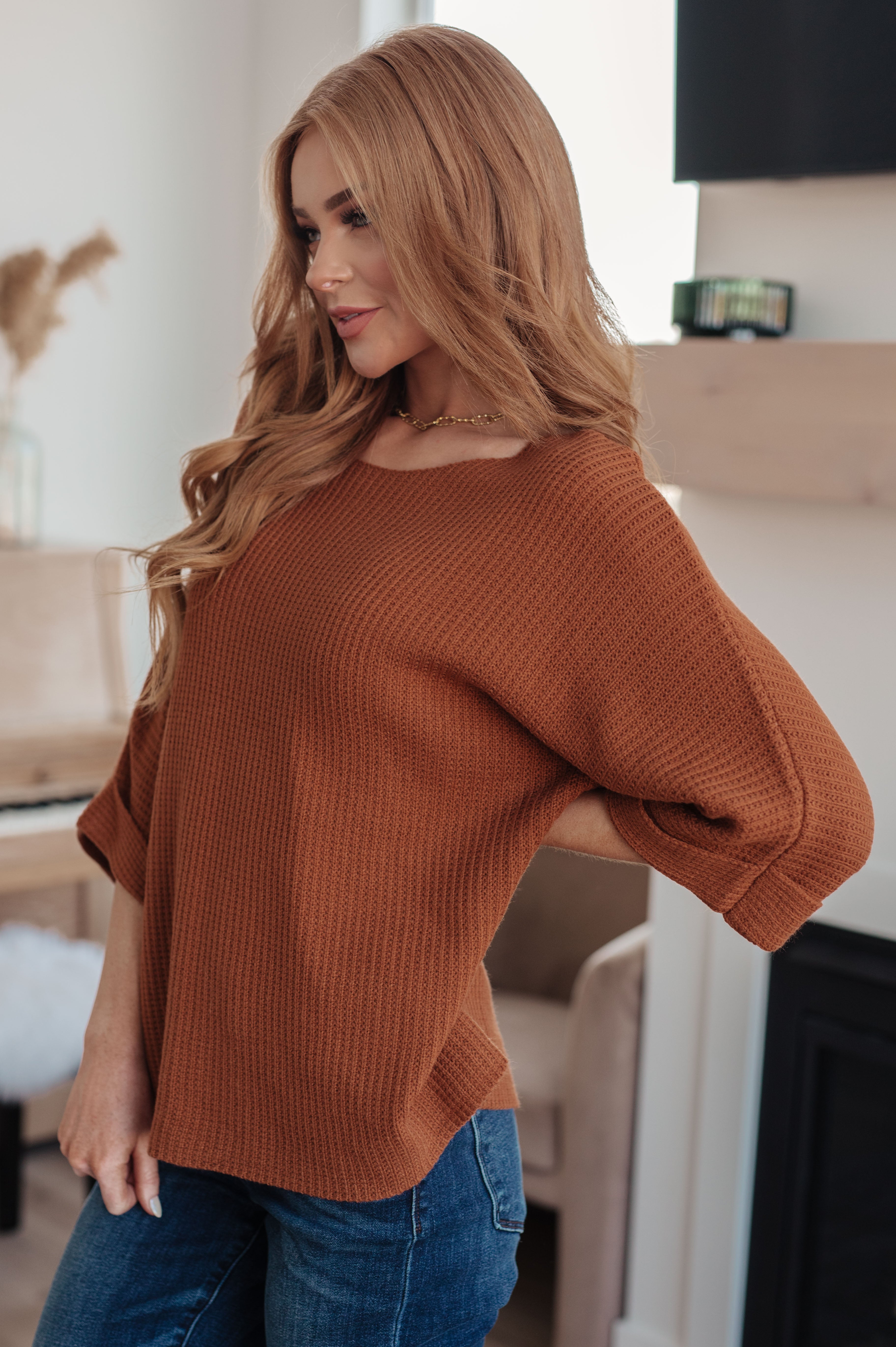 Woman indoors wearing a rust-colored waffle knit sweater with rolled sleeves and dark blue jeans standing near fireplace