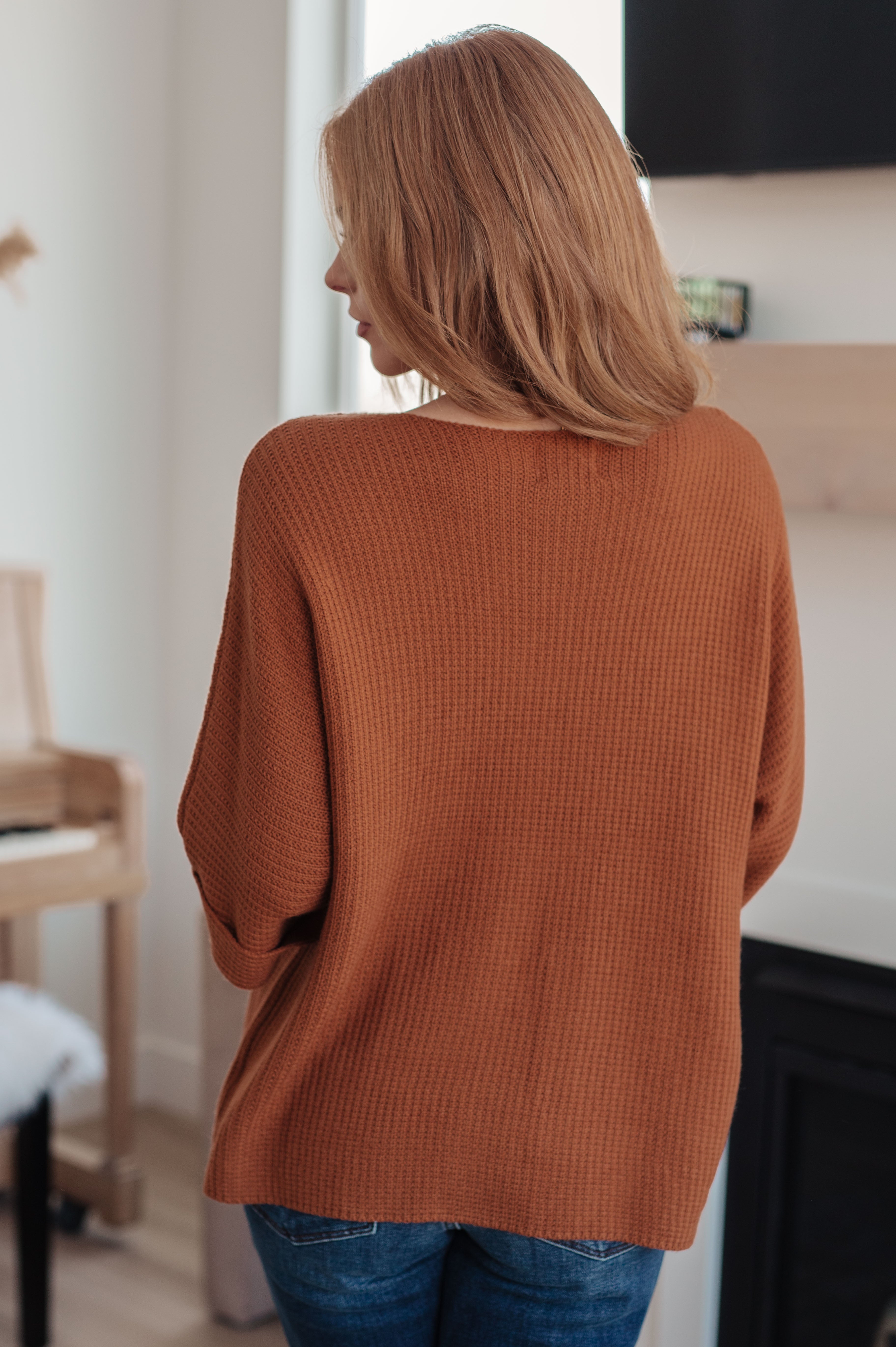 Woman seen from behind wearing a relaxed rust-colored knitted sweater top indoors near piano and fireplace