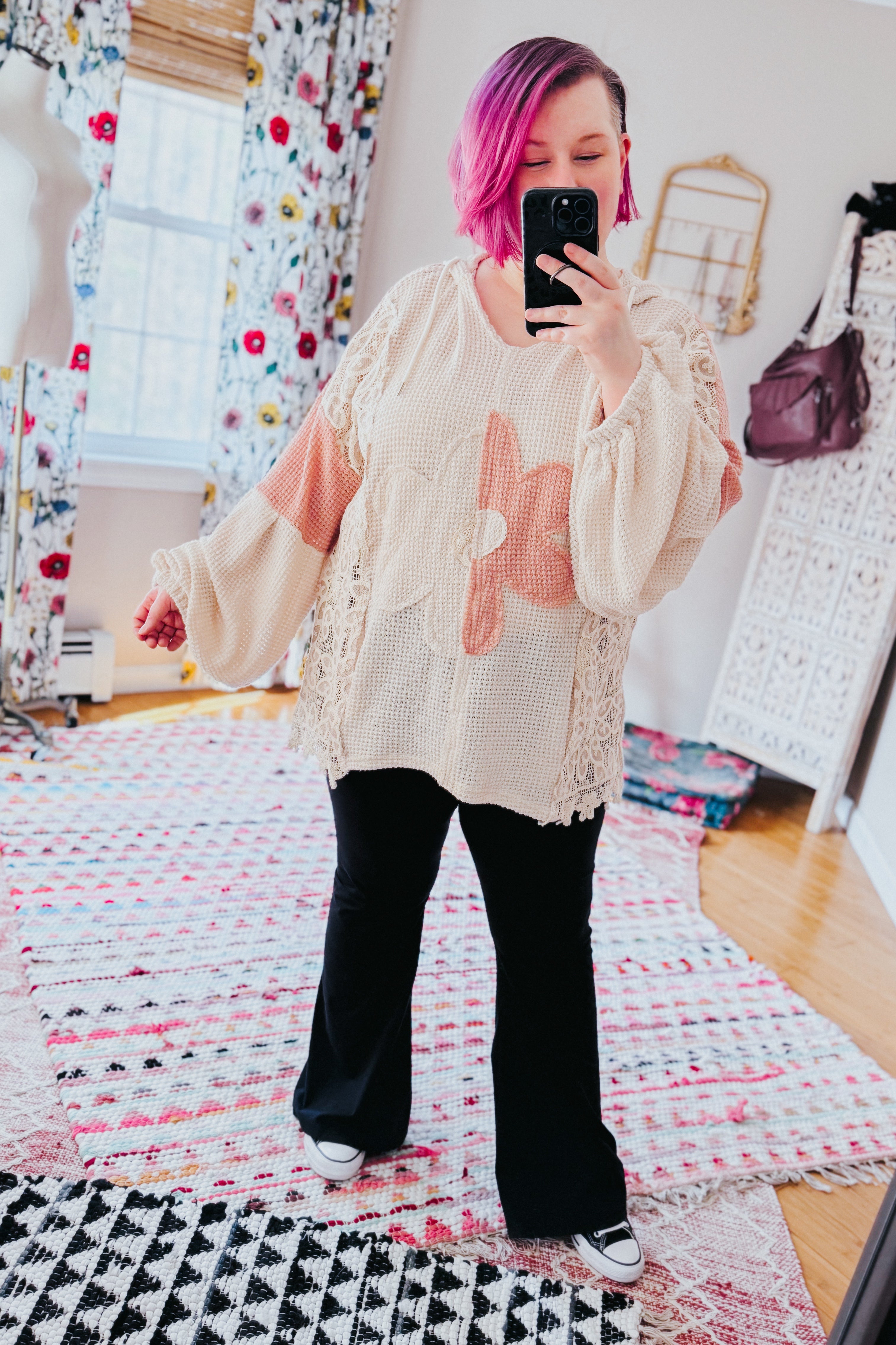 Woman with pink hair wearing a cream textured knit top with large peach floral design and black pants taking selfie