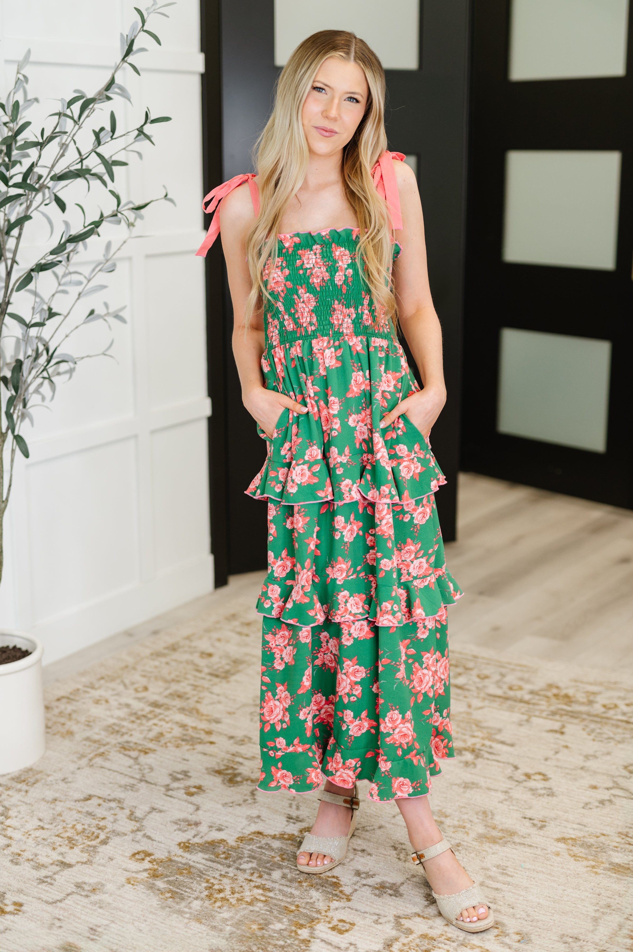 Woman wearing green dress with coral floral print and tiered ruffles standing indoors with hands in pockets