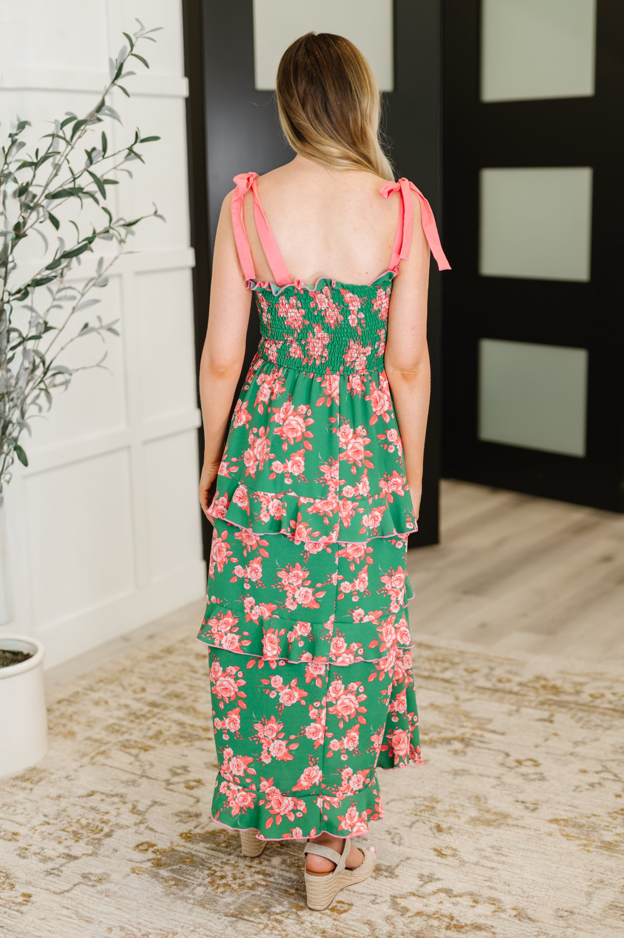 Woman wearing green dress with coral floral print and tiered ruffles with pink tie shoulder straps shown from back indoors