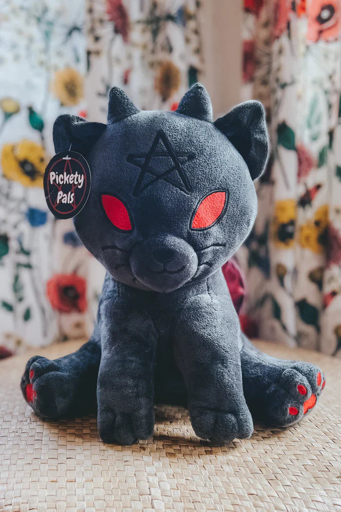 Charcoal black witchy kitten plush toy with red eyes pointed ears and a pentagram detail sitting upright on a woven surface