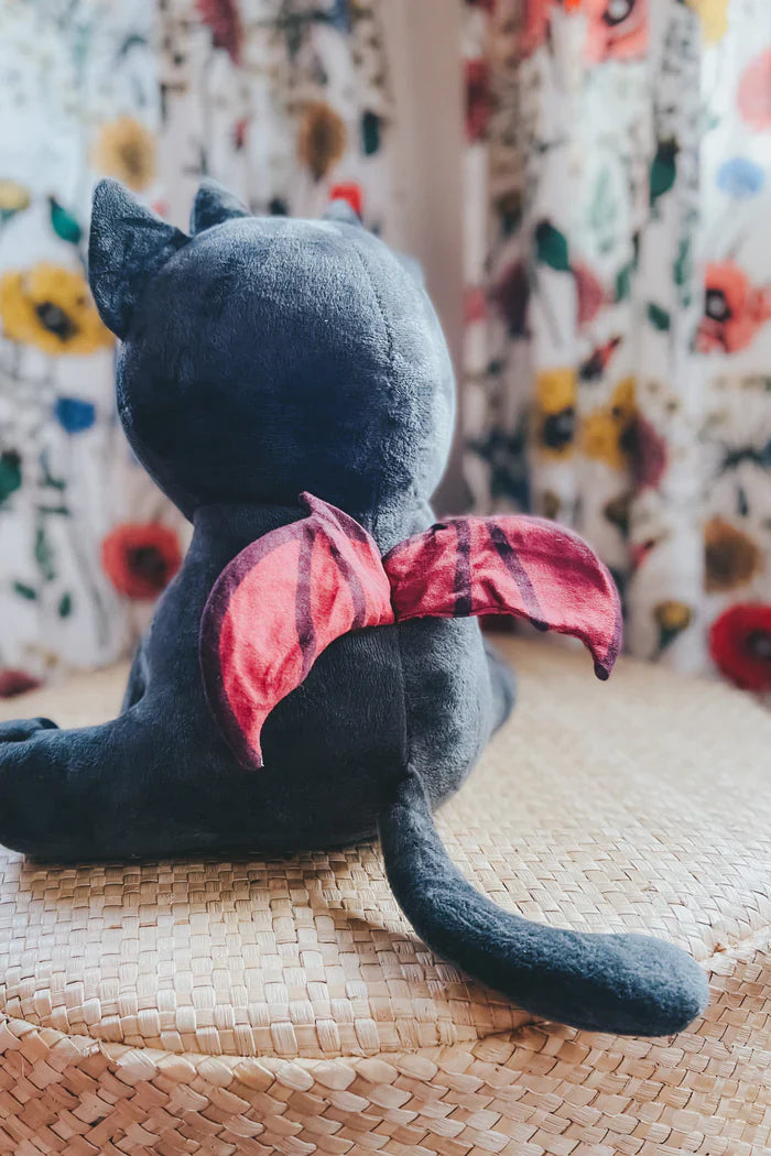 Back view of a charcoal black kitten plush toy with pointed ears long tail and red plaid bow sitting on a woven mat