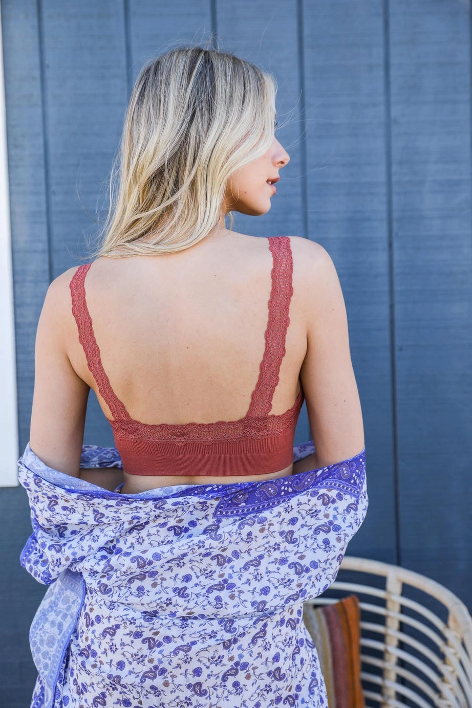 Blonde woman facing away wearing rust lace-trimmed bralette and holding blue floral patterned shawl against gray wall