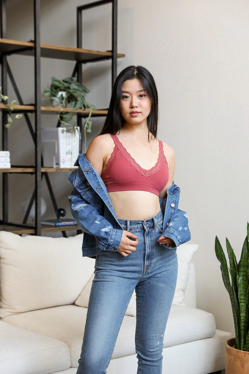 Young woman wearing rust lace-trimmed bralette under denim jacket and high-waisted jeans indoors by shelves and sofa