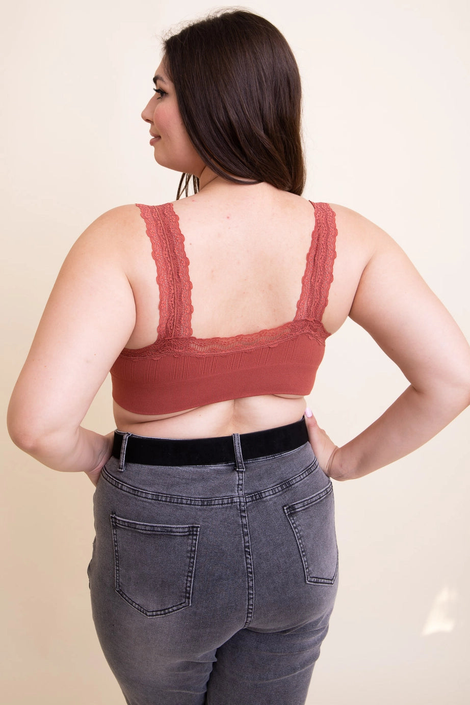 Woman with dark hair wearing rust-colored lace-trimmed bralette and high-waisted dark gray jeans shown from back view