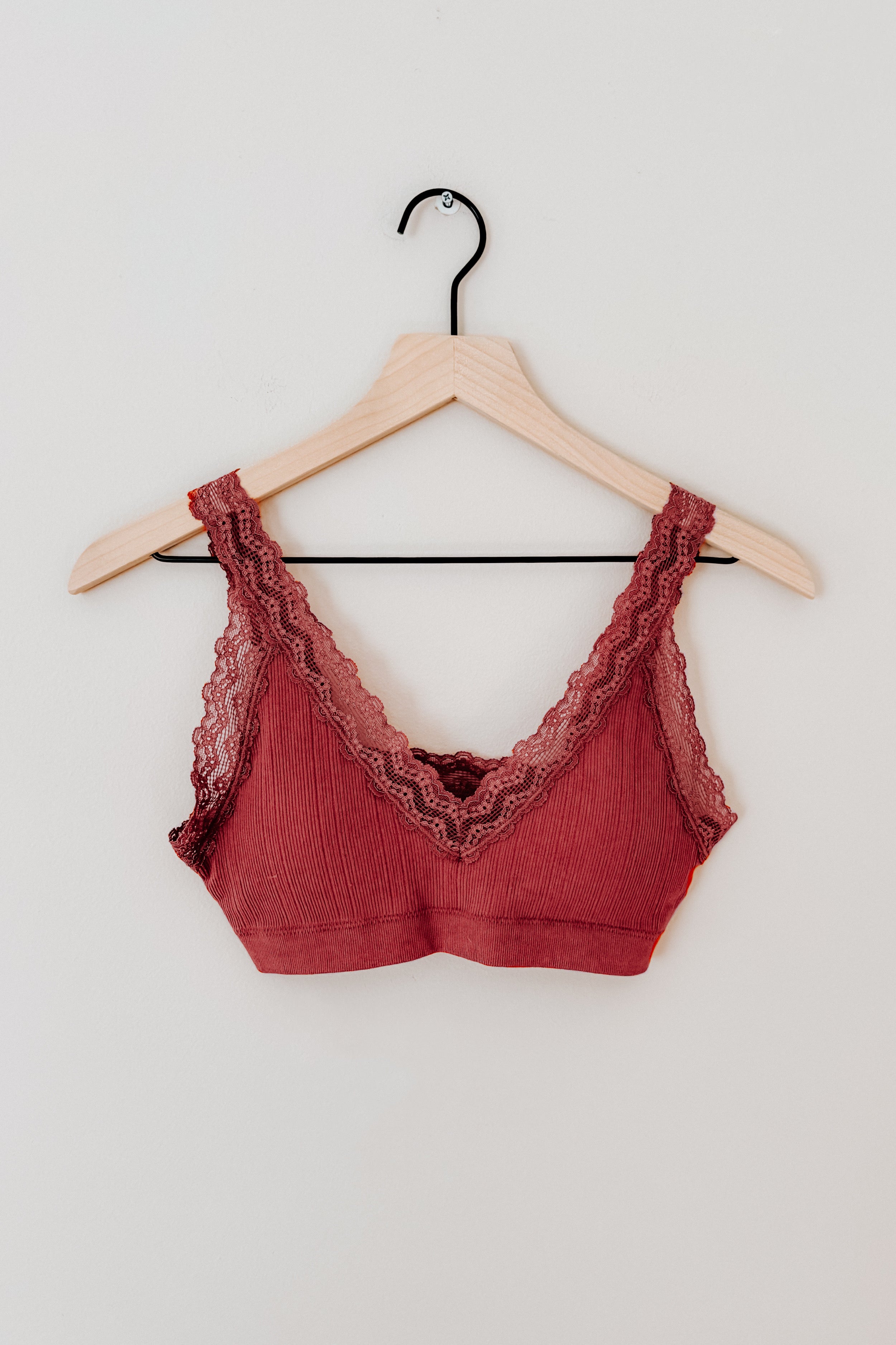 Rust-colored ribbed bralette with scalloped lace trim hanging on wooden hanger against plain wall background