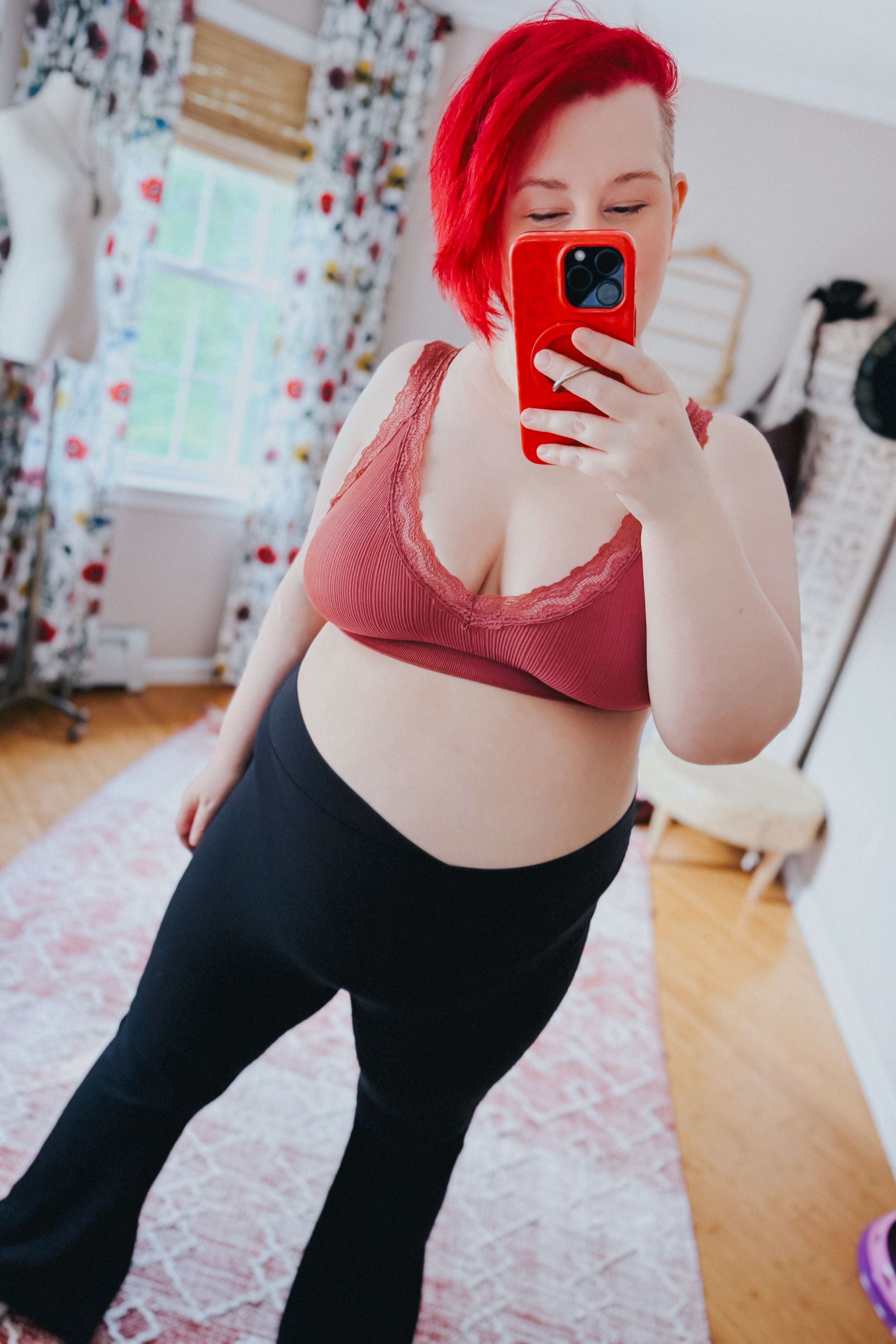 Woman with bright red hair wearing rust-colored lace-trimmed bralette and black pants taking selfie in cozy room