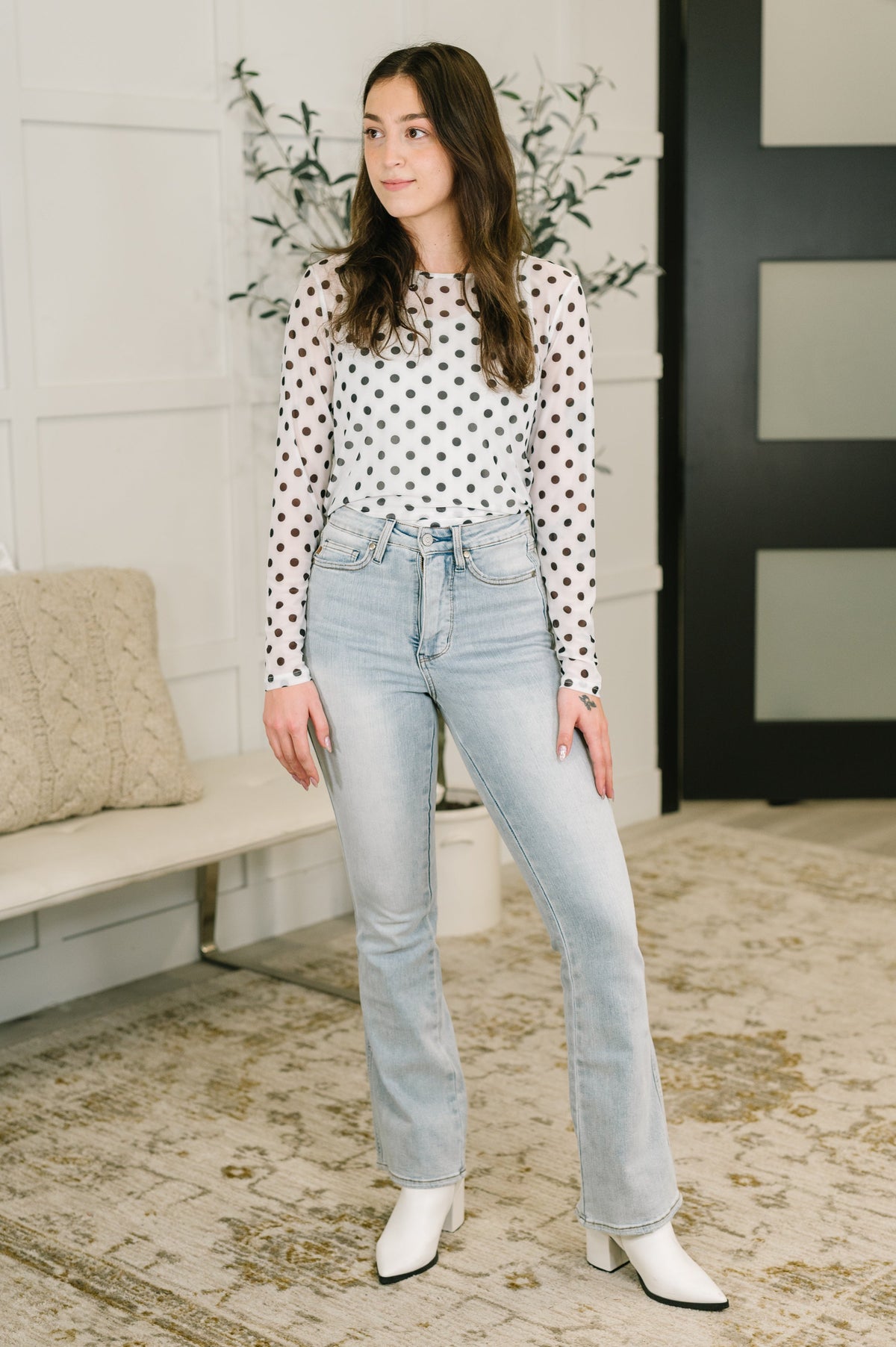 Woman wearing light wash high waist bootcut jeans with tummy control styled with a white polka dot blouse and white boots indoors