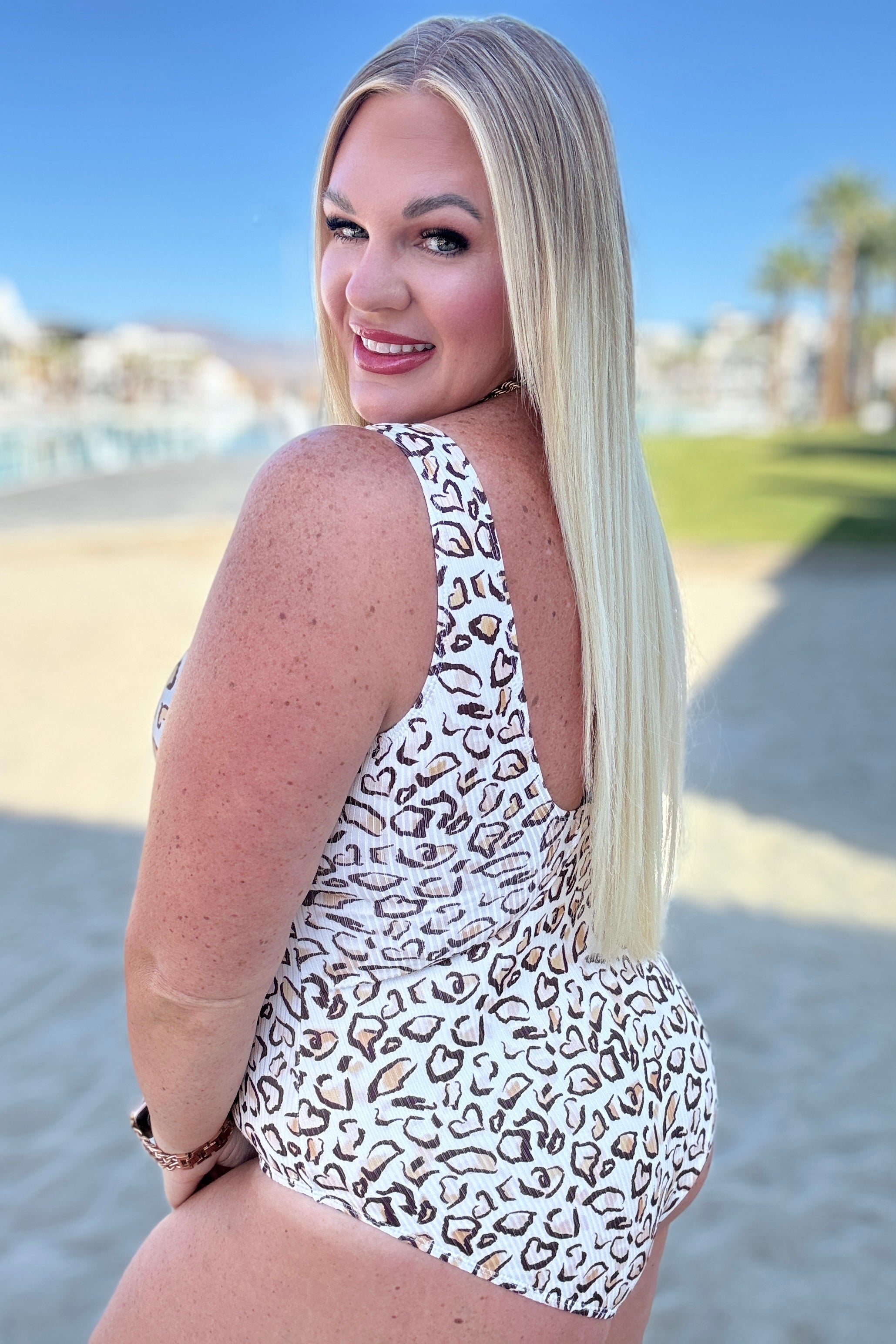 Woman with long blonde hair wearing a white and mocha leopard print swimsuit smiling outdoors near beach area