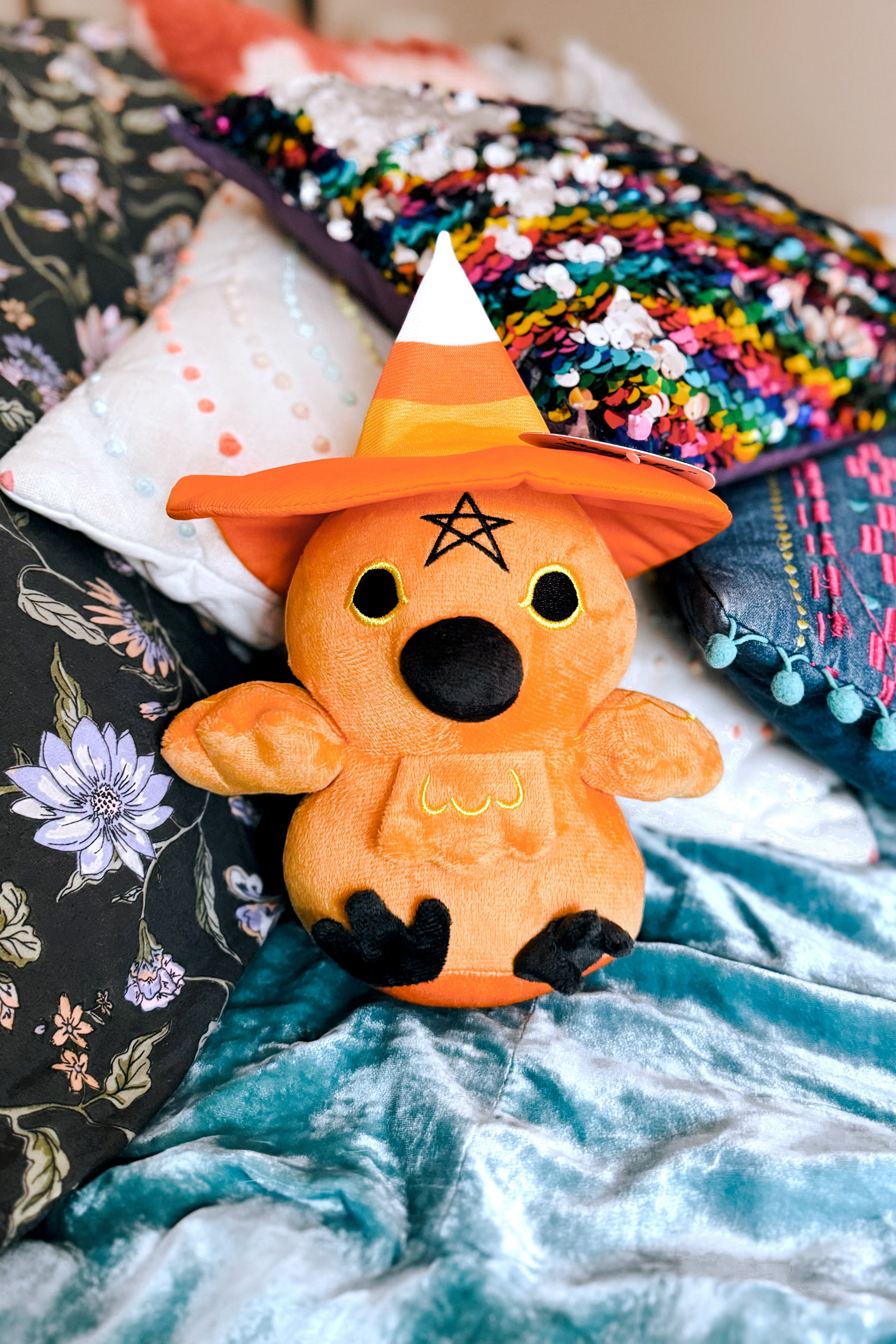 Plush orange bird wearing a pumpkin spice colored witch hat sitting on a bed with colorful pillows and blankets