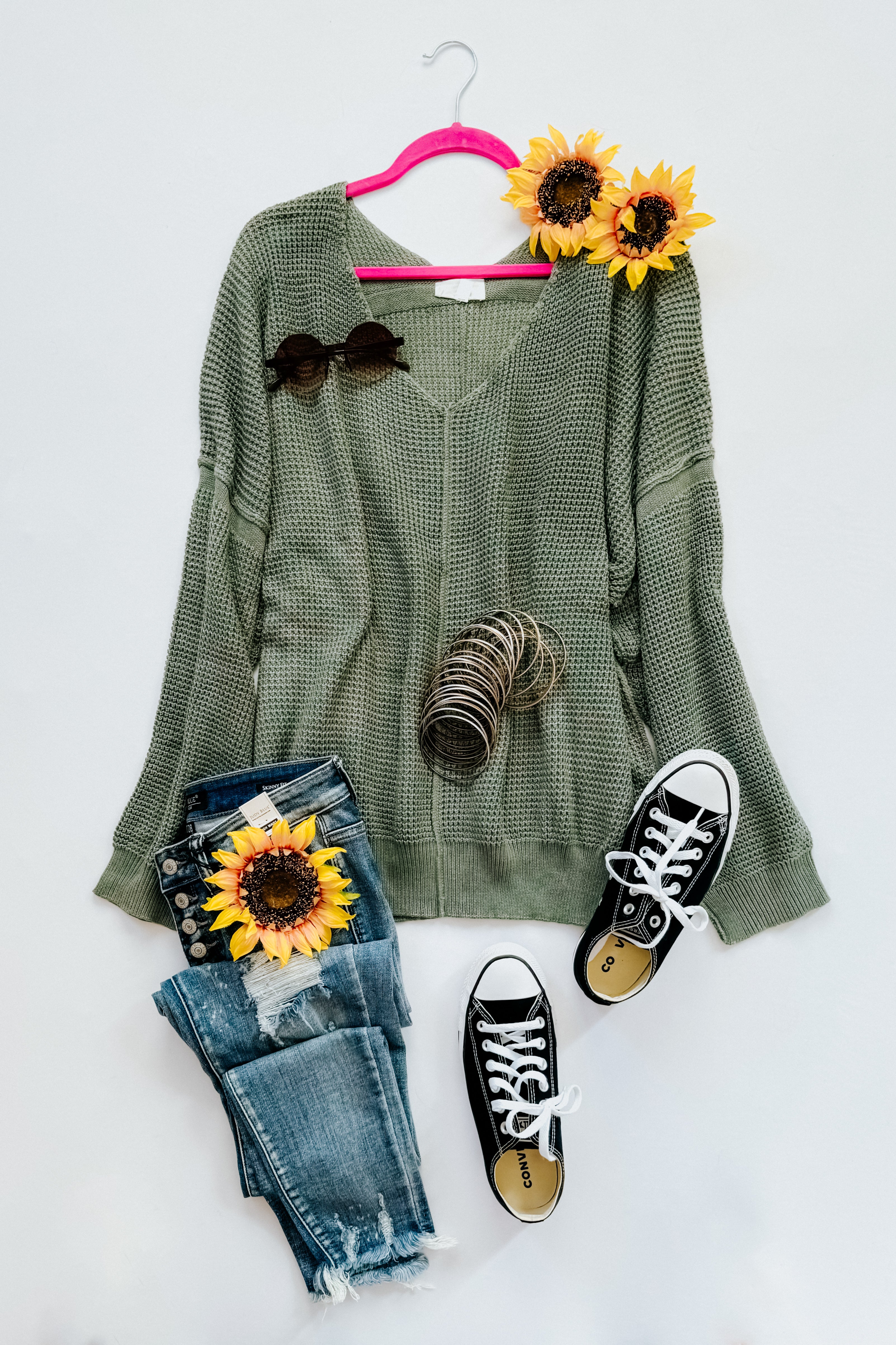 A flat-lay photo with an olive green v-neck sweater with exposed seams, jeans, converse sneakers, accessories and floral elements on a white background.