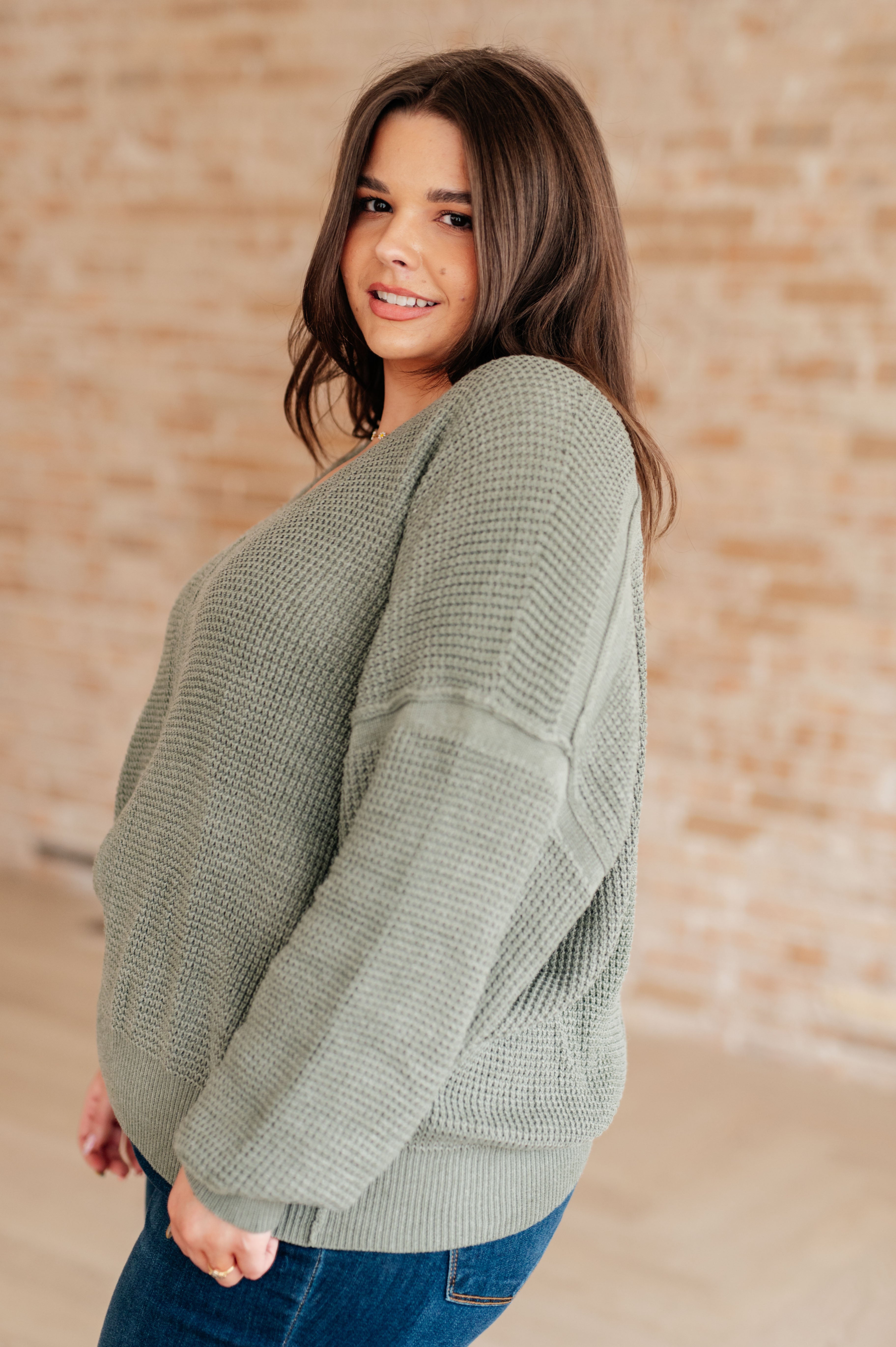 Woman wearing an olive green v-neck sweater with exposed seams and blue jeans standing against a brick wall