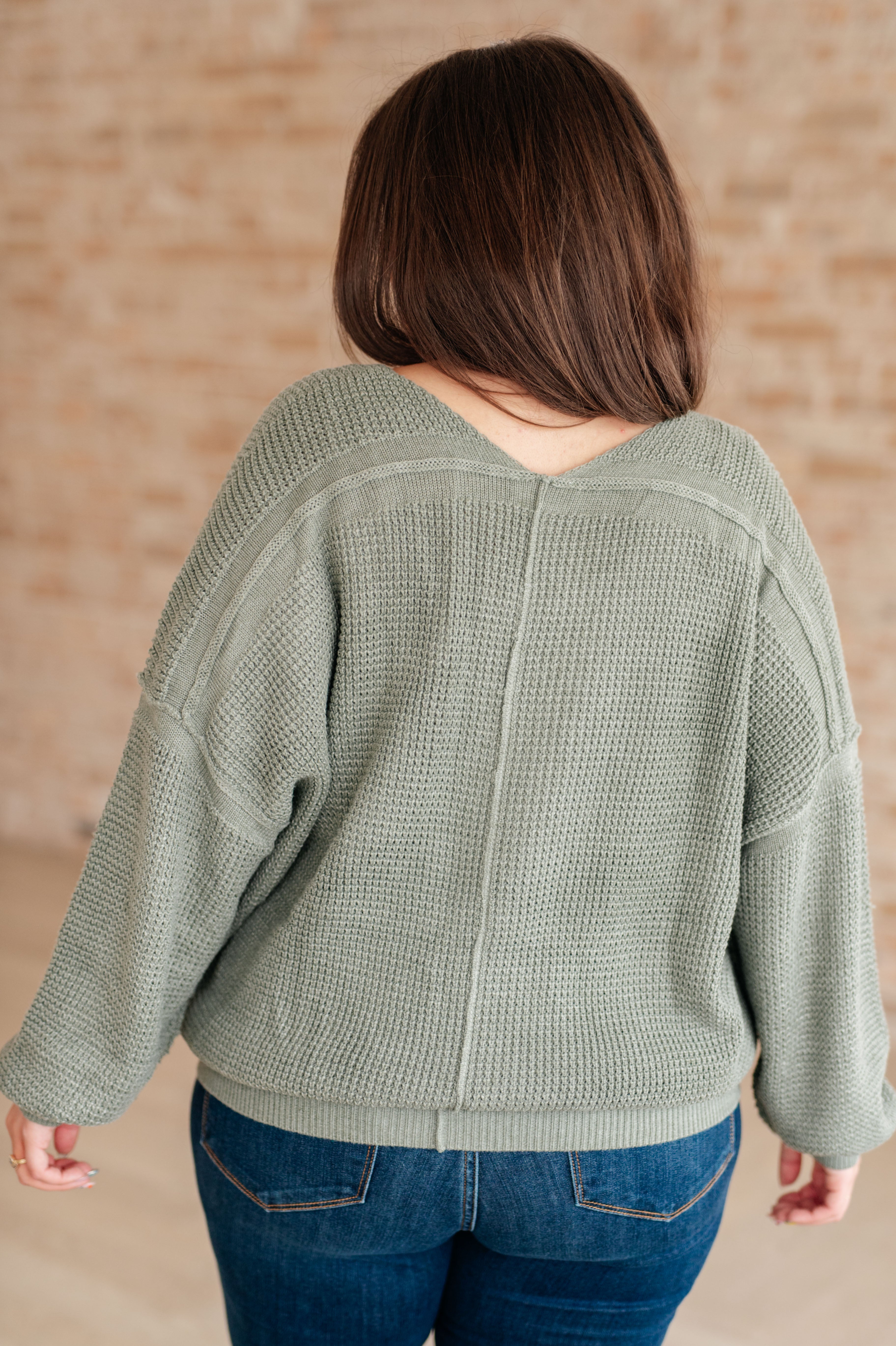 Woman wearing an olive green v-neck sweater with exposed seams and blue jeans standing against a brick wall
