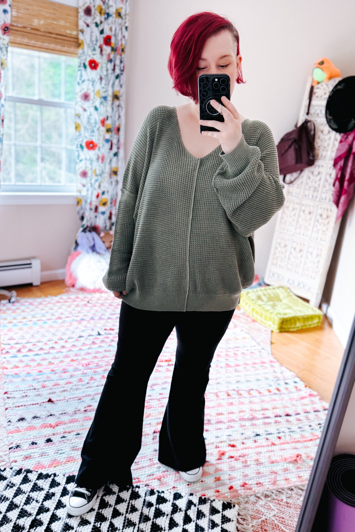 Red haired woman modeling a sage V neck sweater with exposed seams black flared pants and white sneakers in a colorful bedroom rug setting