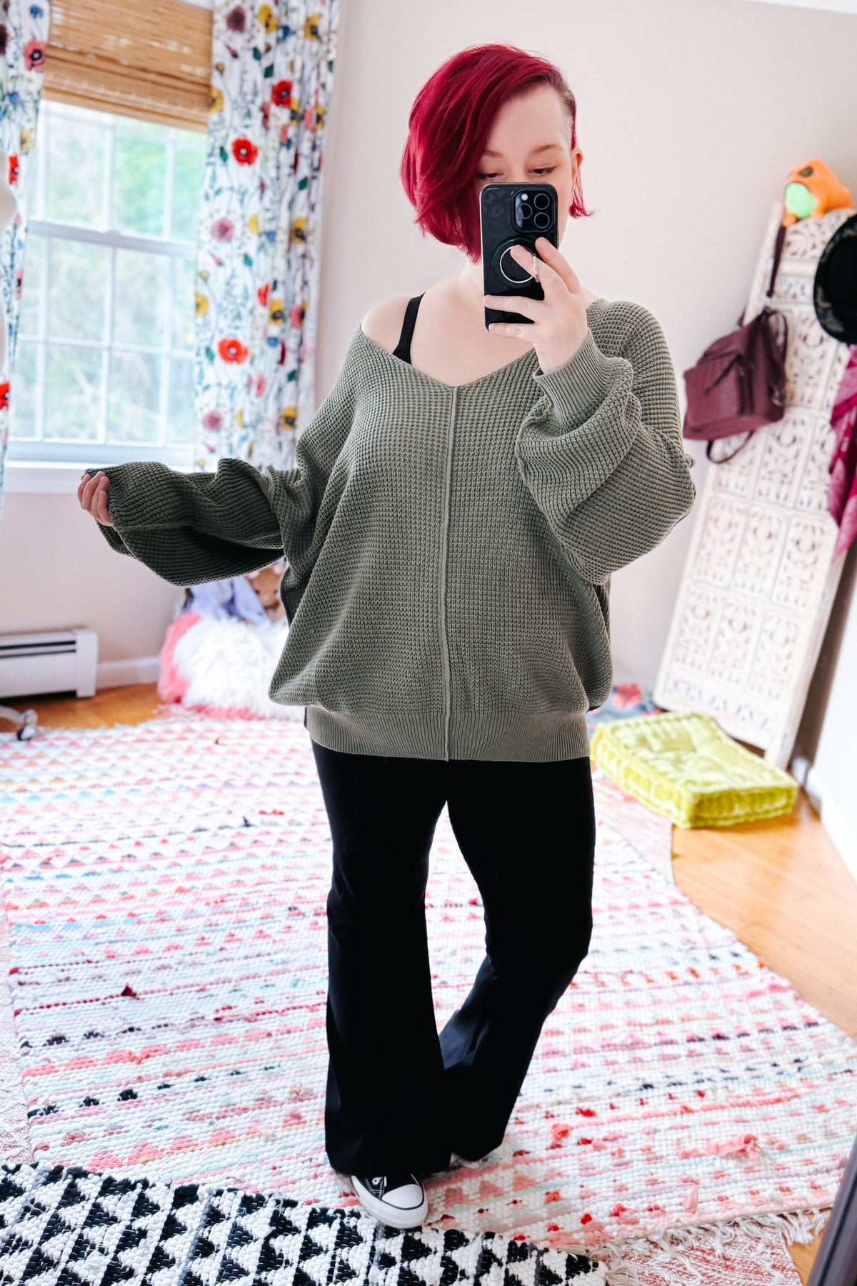 Red haired woman taking a bedroom mirror selfie in a sage V neck sweater with black pants and white sneakers on a patterned rug