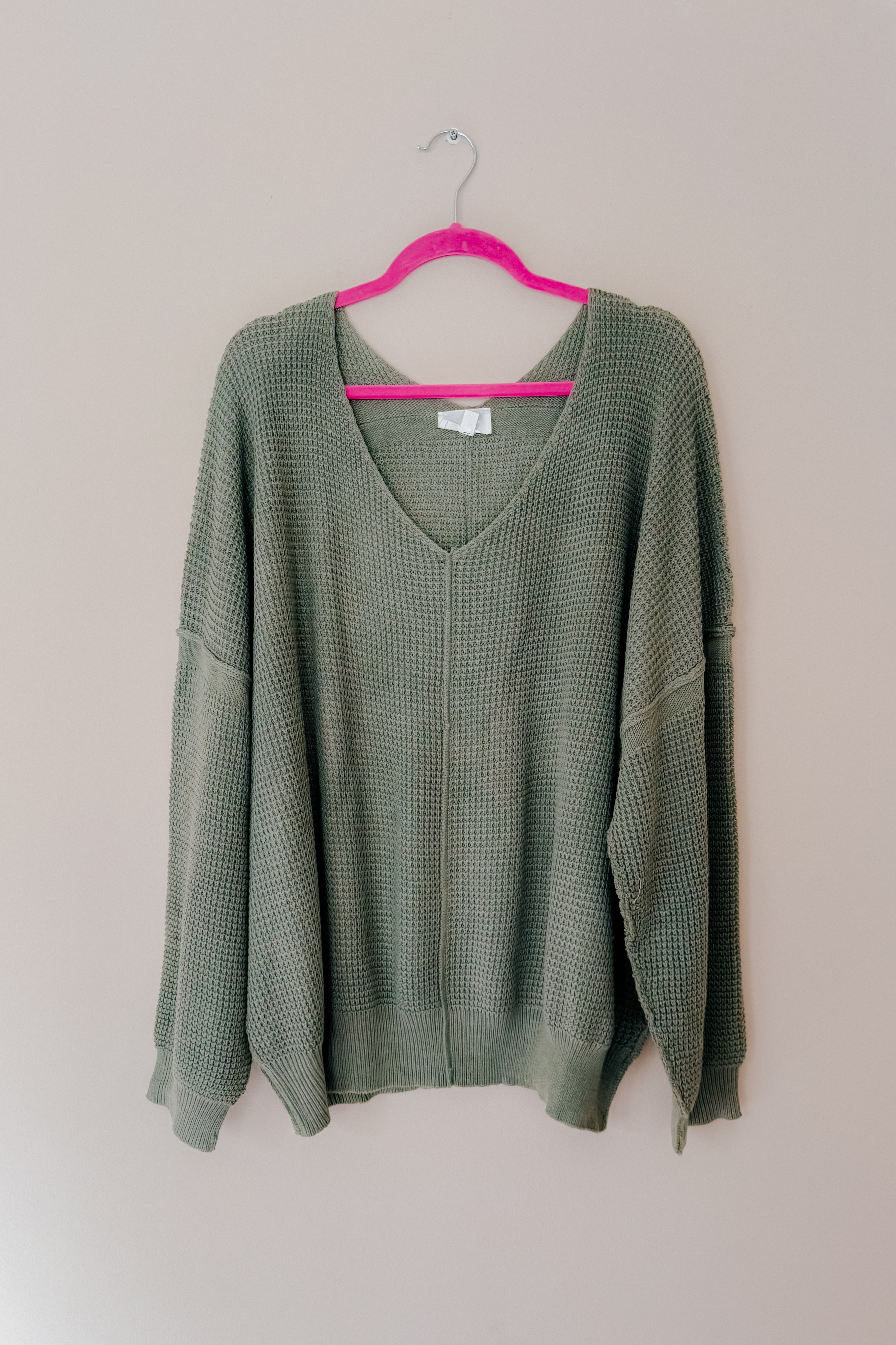 A olive green v-neck sweater with exposed seams hanging on a pink hanger.
