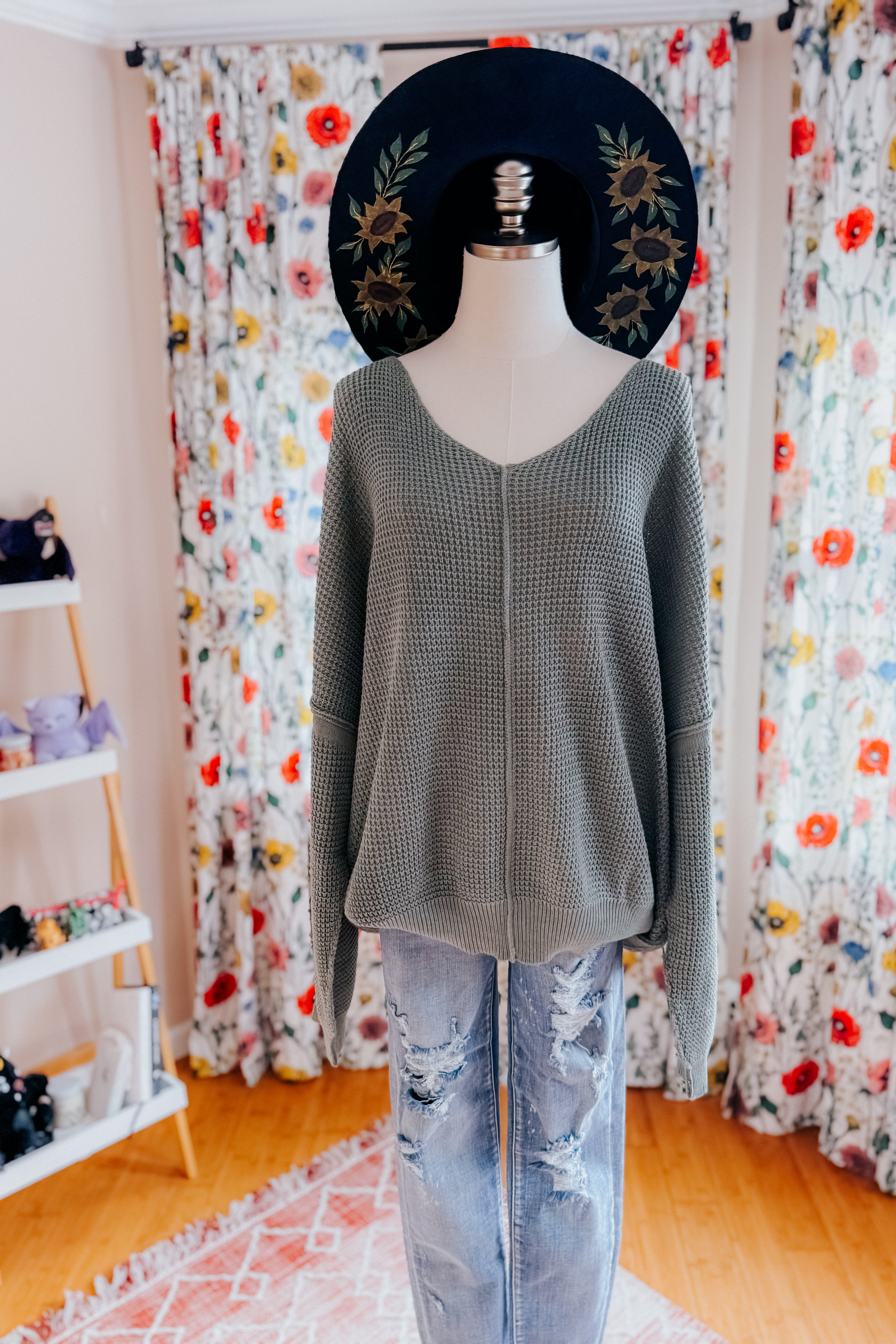 A dress form wearing an olive green v-neck sweater with exposed seams, a hand painted wide brim hat, and distressed blue jeans in an indoor setting.