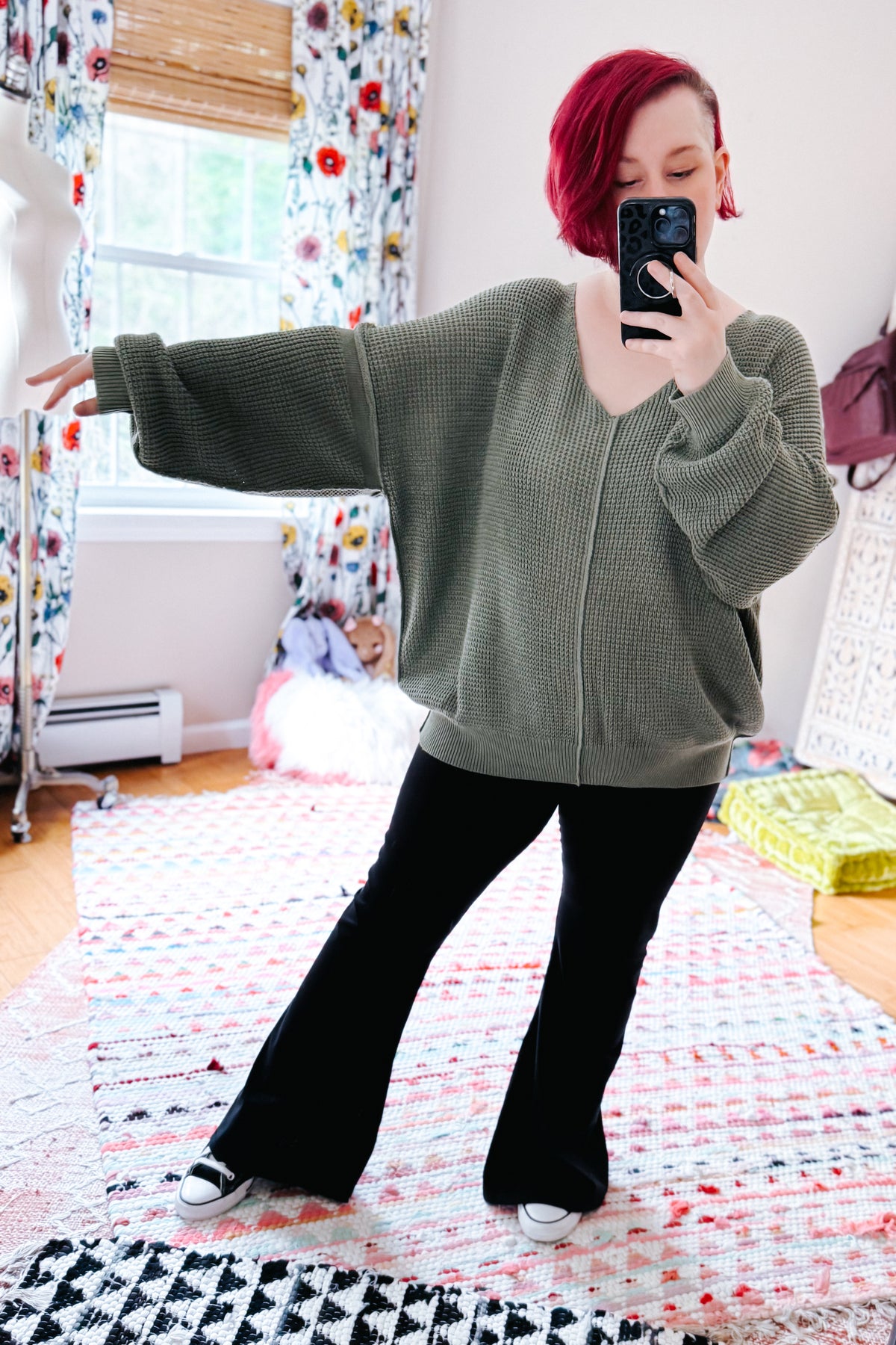 Red haired woman posing in a sage V neck sweater with exposed seams black flared pants and sneakers during a bedroom mirror selfie