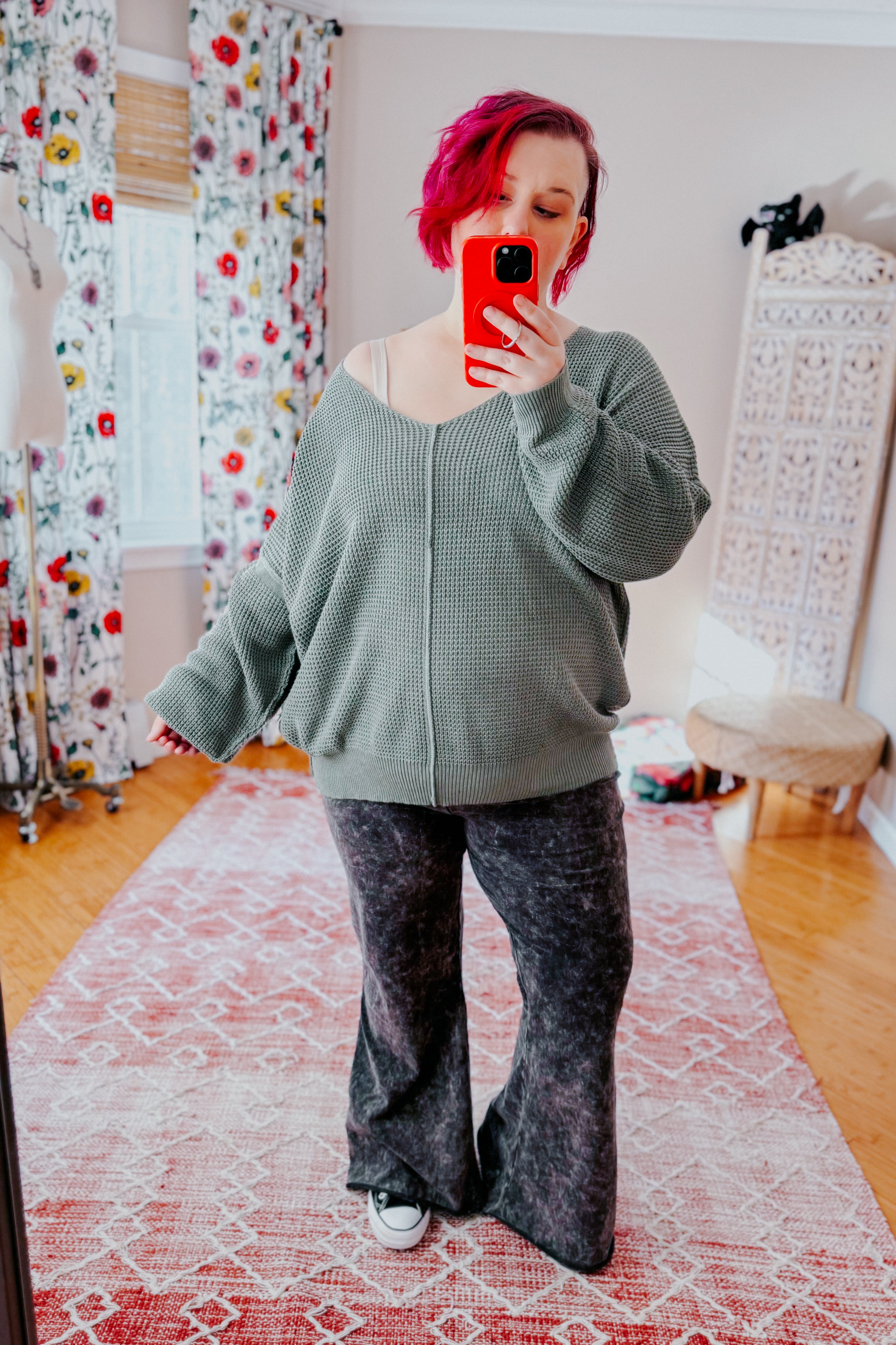 A women with red hair taking a mirror selfie wearing an olive green v-neck sweater with exposed seams, grey acid wash pants, and converse sneakers standing in an indoor setting.