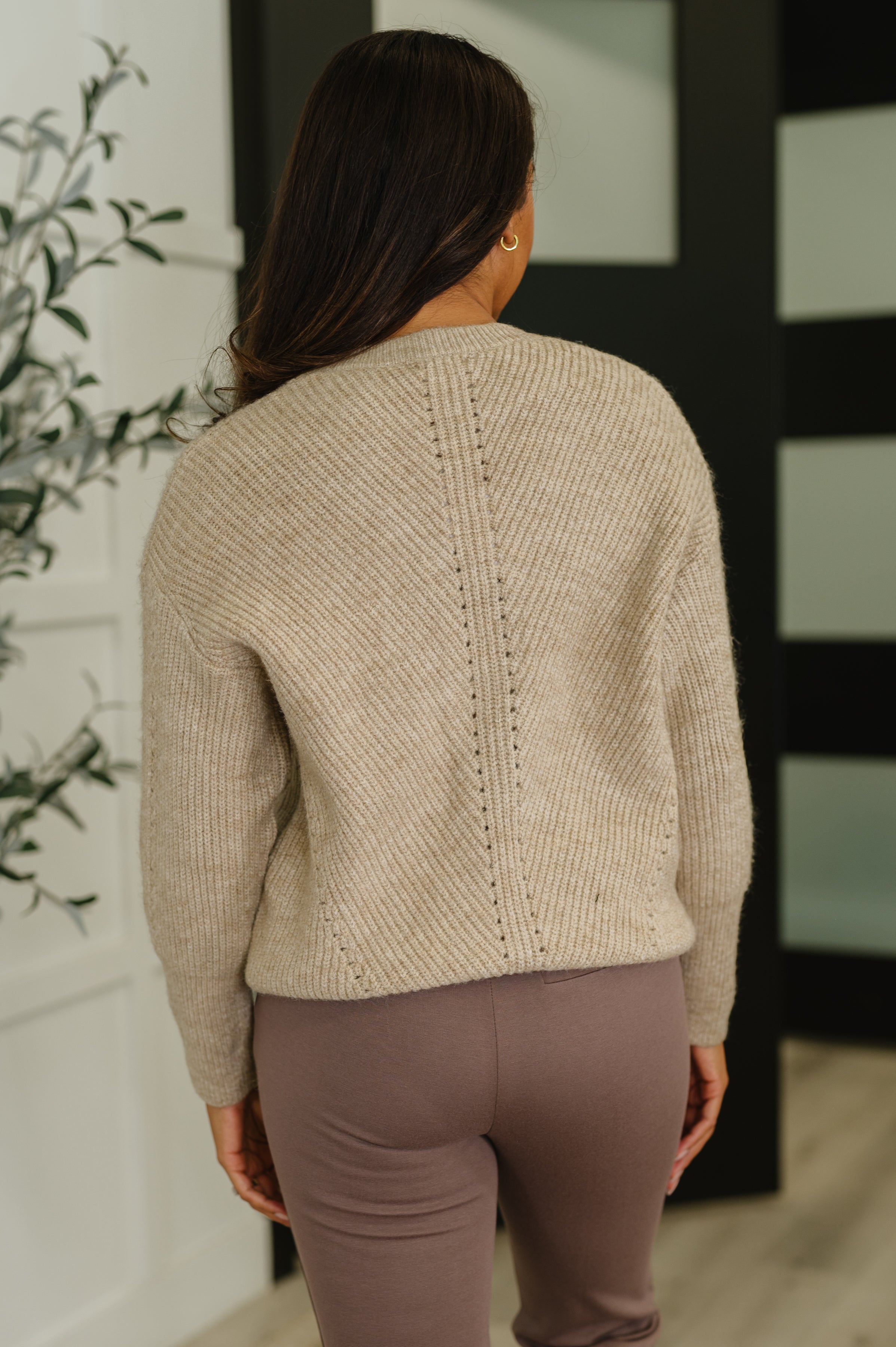 Woman wearing an oatmeal ribbed knit sweater with vertical stitch details paired with mauve fitted pants indoors