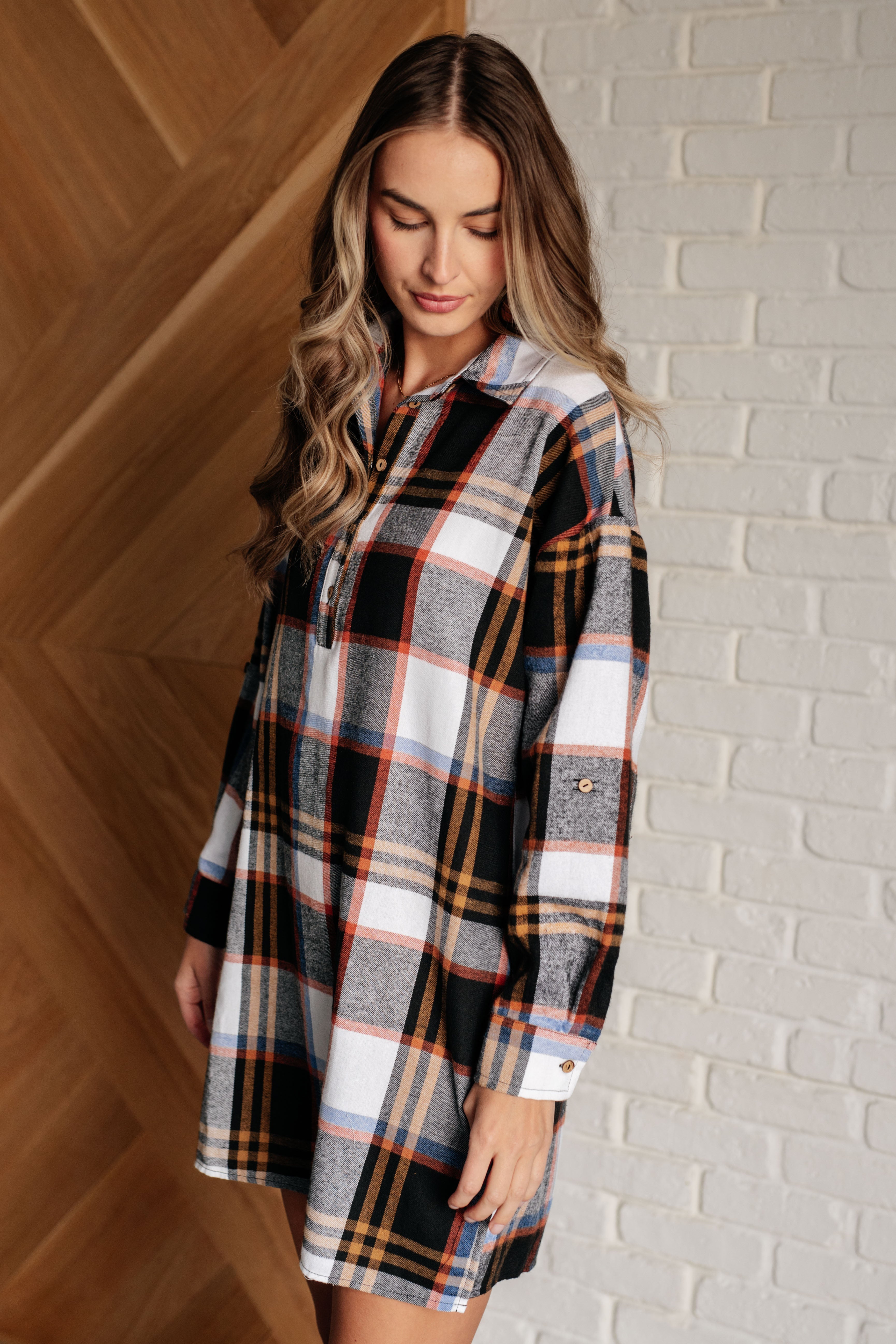 Woman wearing a black plaid shirt dress with orange, beige, and blue accents standing indoors by brick and wood walls