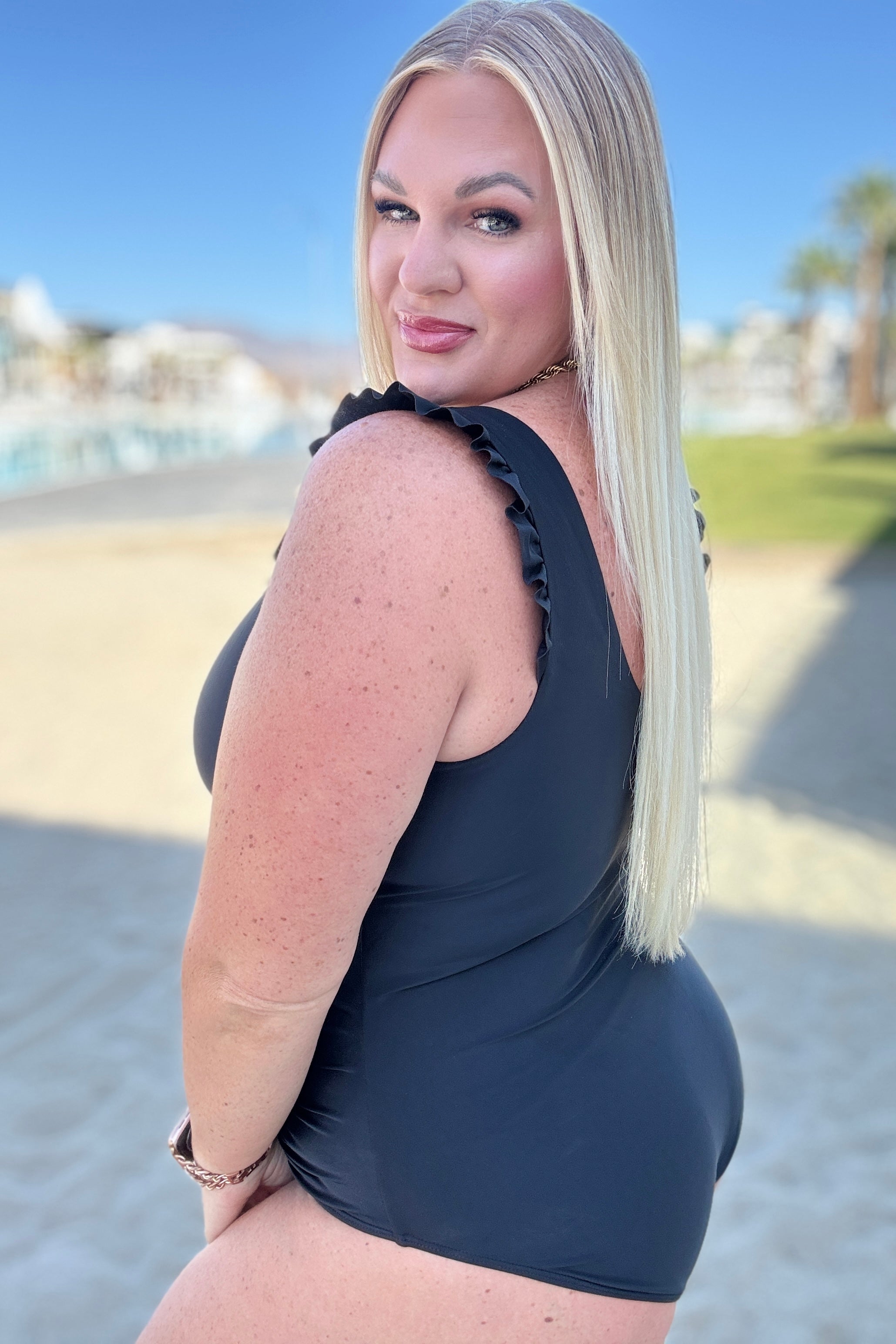 Blonde woman posing outdoors wearing a black swimsuit with ruffled shoulder straps on a sunny day