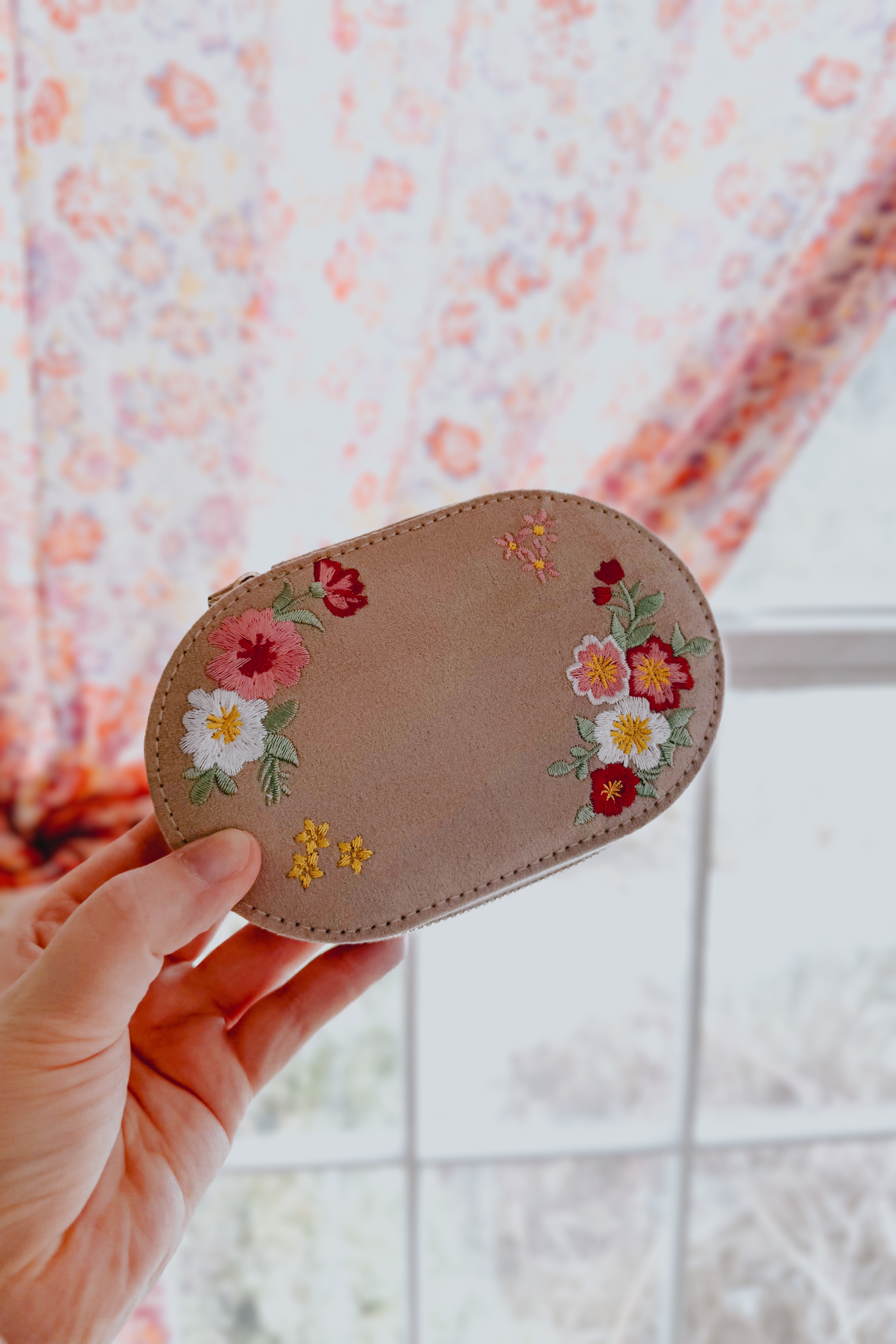 Hand holding a small cream velvet case with colorful floral embroidery against blurred window background
