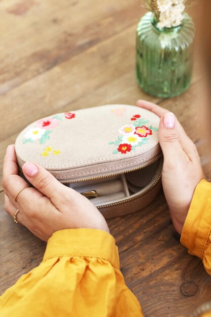 Hands holding a cream velvet jewelry box with floral embroidery and gold zipper on a wooden surface