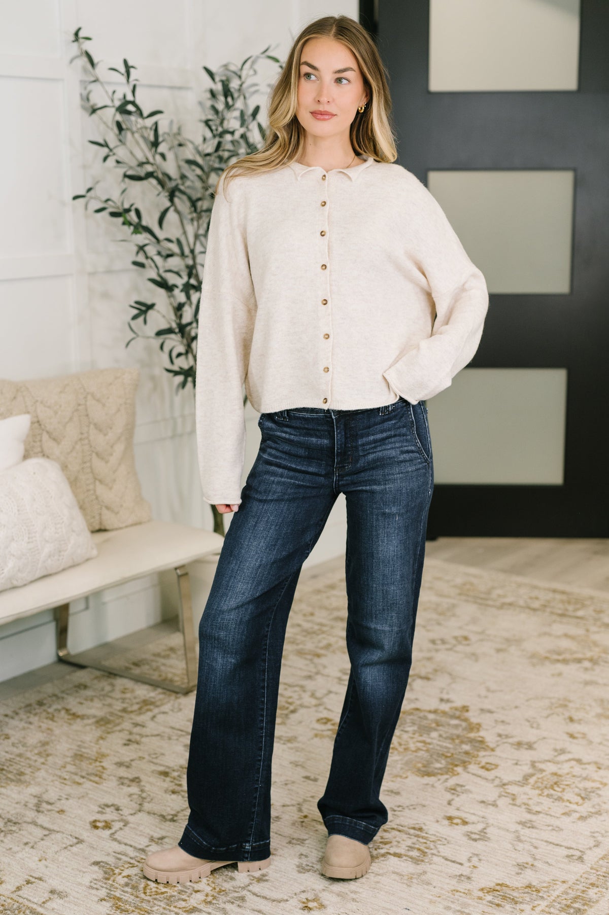Woman modeling dark blue high waist wide leg jeans with a cream button front cardigan in a cozy modern home interior