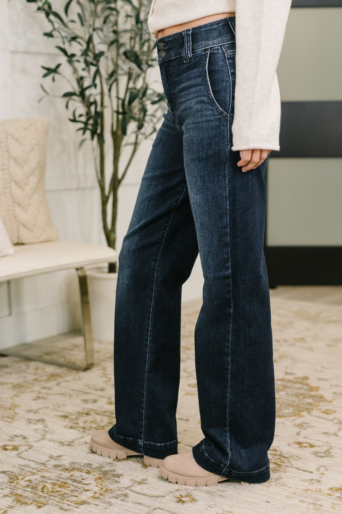 Woman wearing dark wash blue high waist wide leg jeans with a cream sweater and neutral sneakers in a modern home interior
