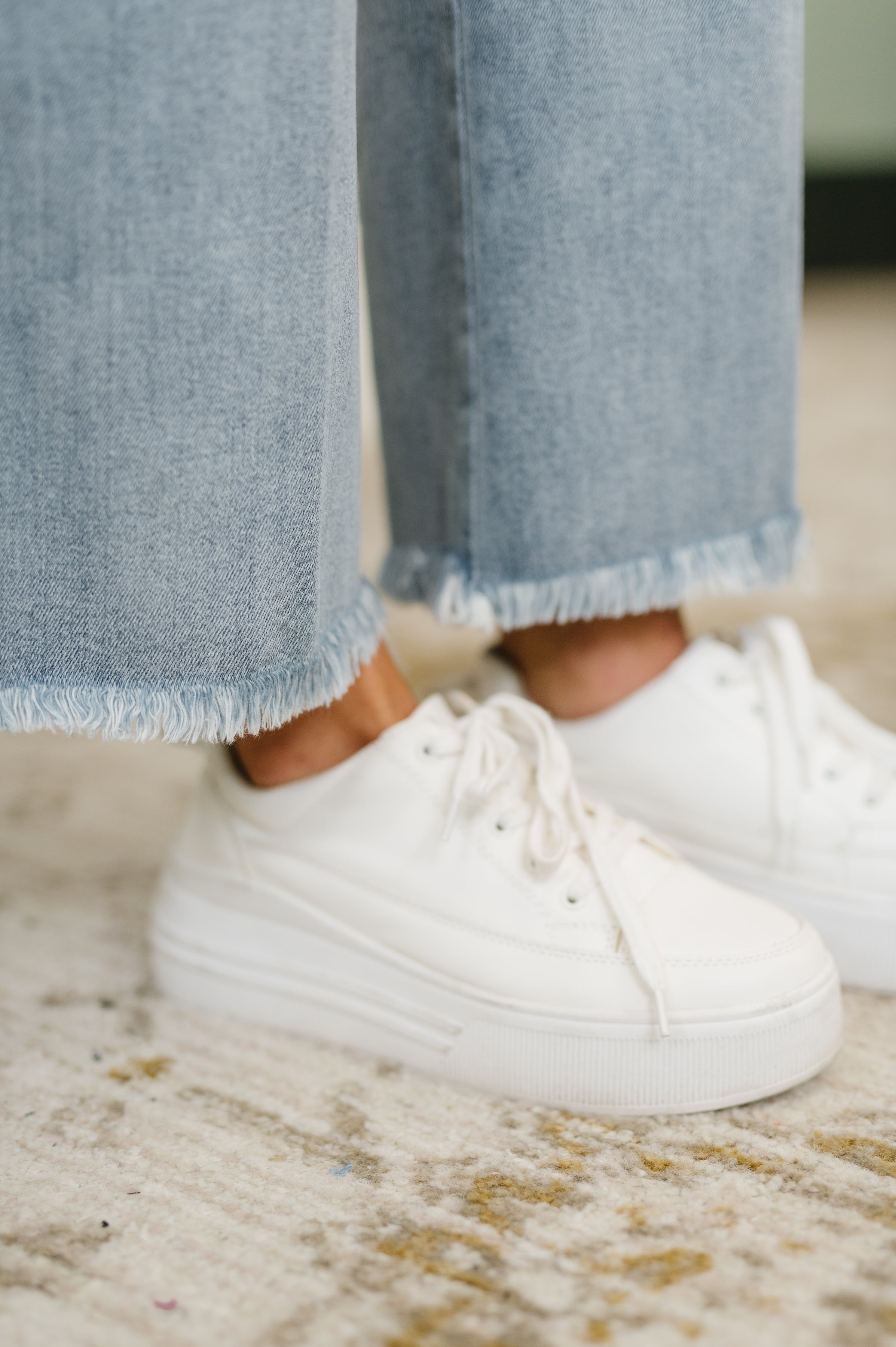 Close-up of light blue denim wide-leg jeans with frayed hems paired with white platform sneakers on textured floor