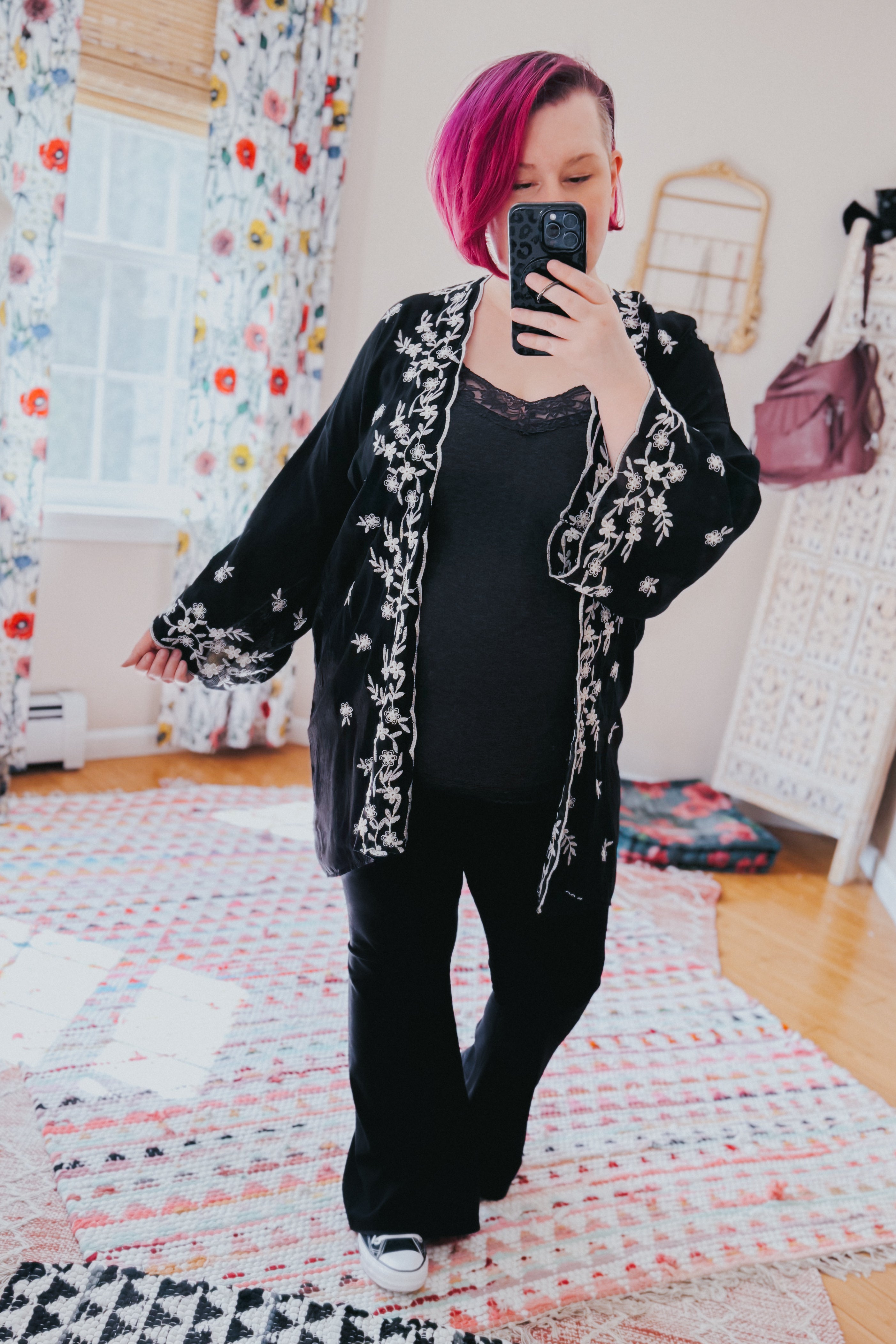 Woman taking mirror selfie wearing a black duster with white floral embroidery over black pants and top indoors