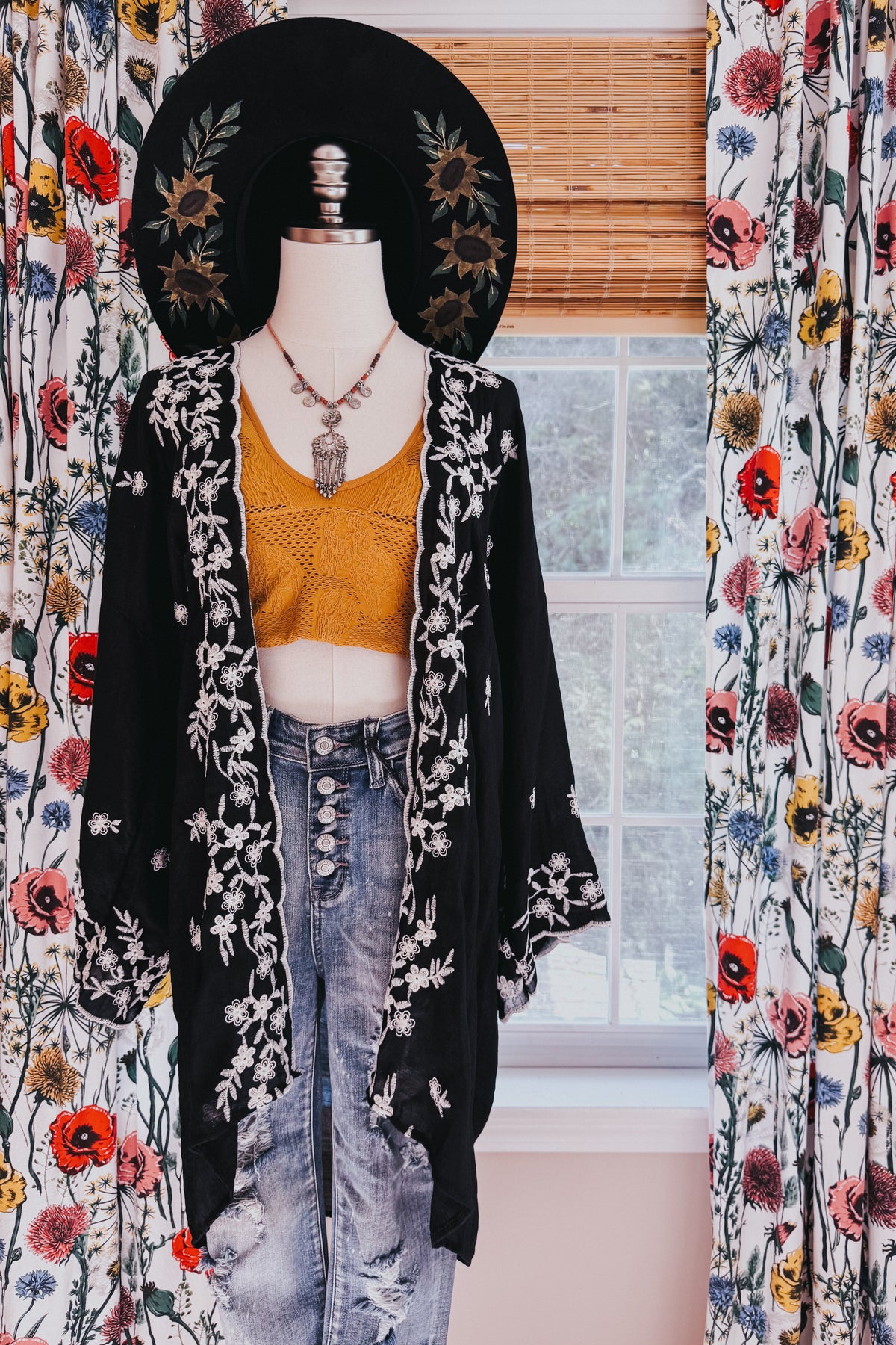 Black floral embroidered duster layered over a mustard knit crop top and distressed jeans on a mannequin in a floral room