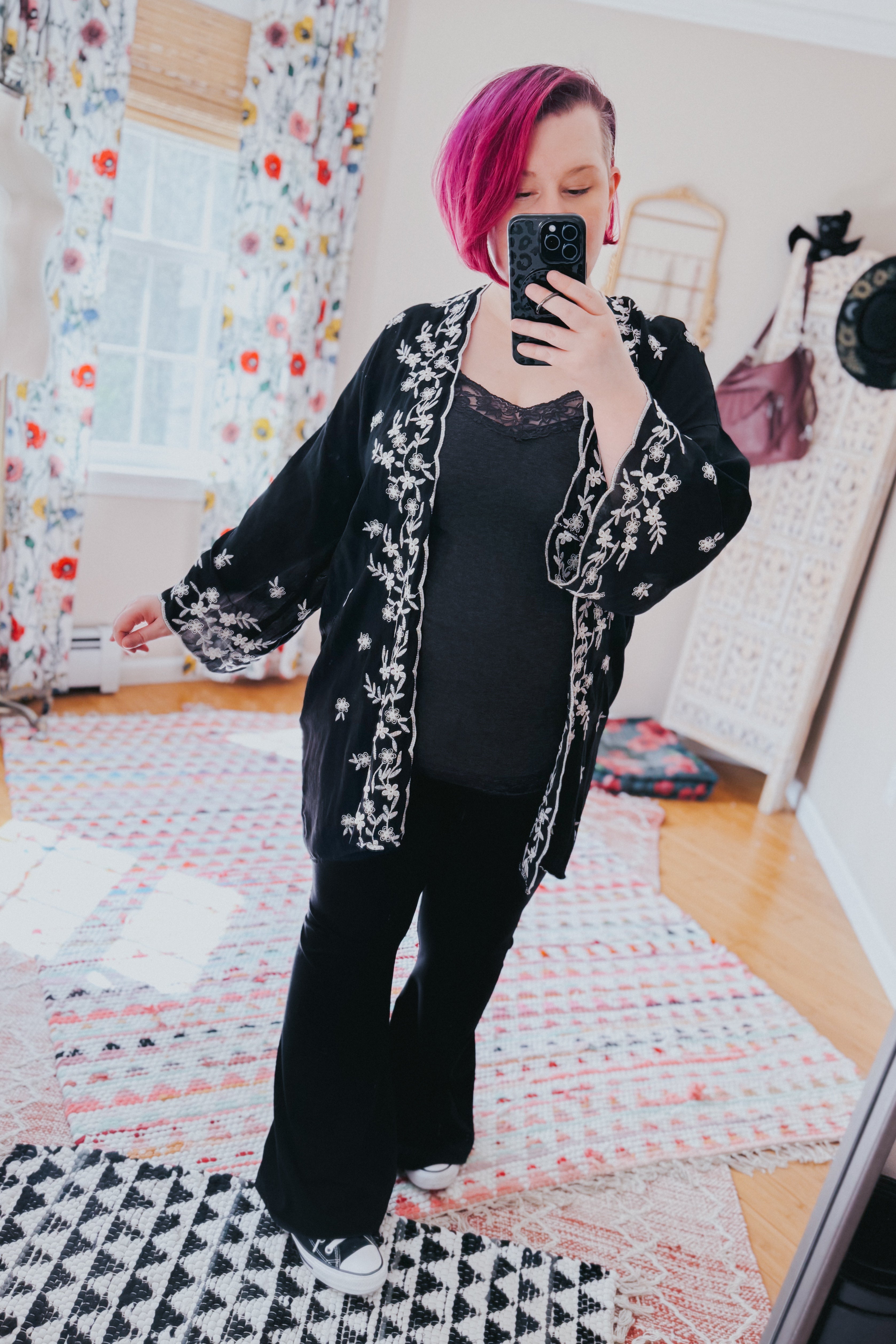 Woman wearing a black duster with white floral embroidery over a dark lace-trimmed top and black pants indoors
