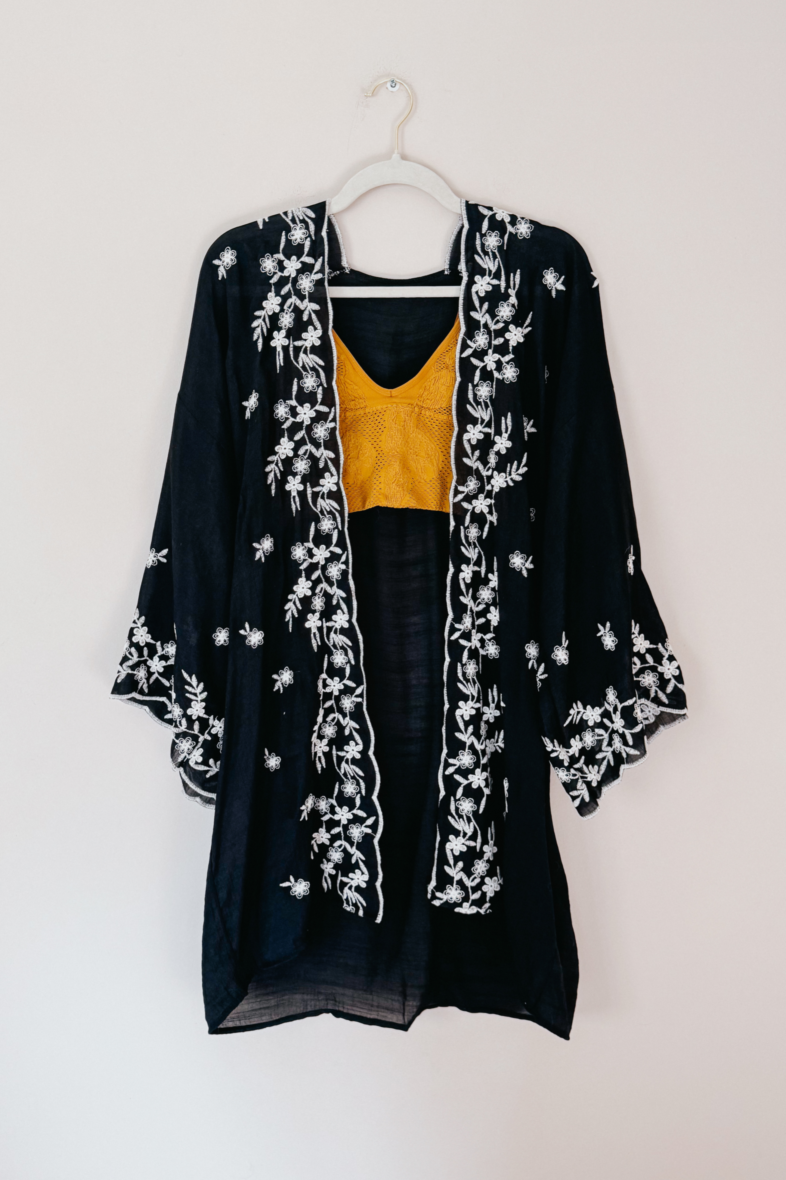 Black semi-sheer duster with white floral embroidery layered over a mustard crochet top hanging on white hanger